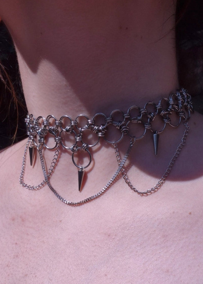 Zbliżenie na szyję z ręcznie robionym chokerem chainmaille ze stali nierdzewnej, ozdobionym kolcami, łańcuszkami i dużymi metalowymi ogniwami. Styl punk, industrial i alternatywny.