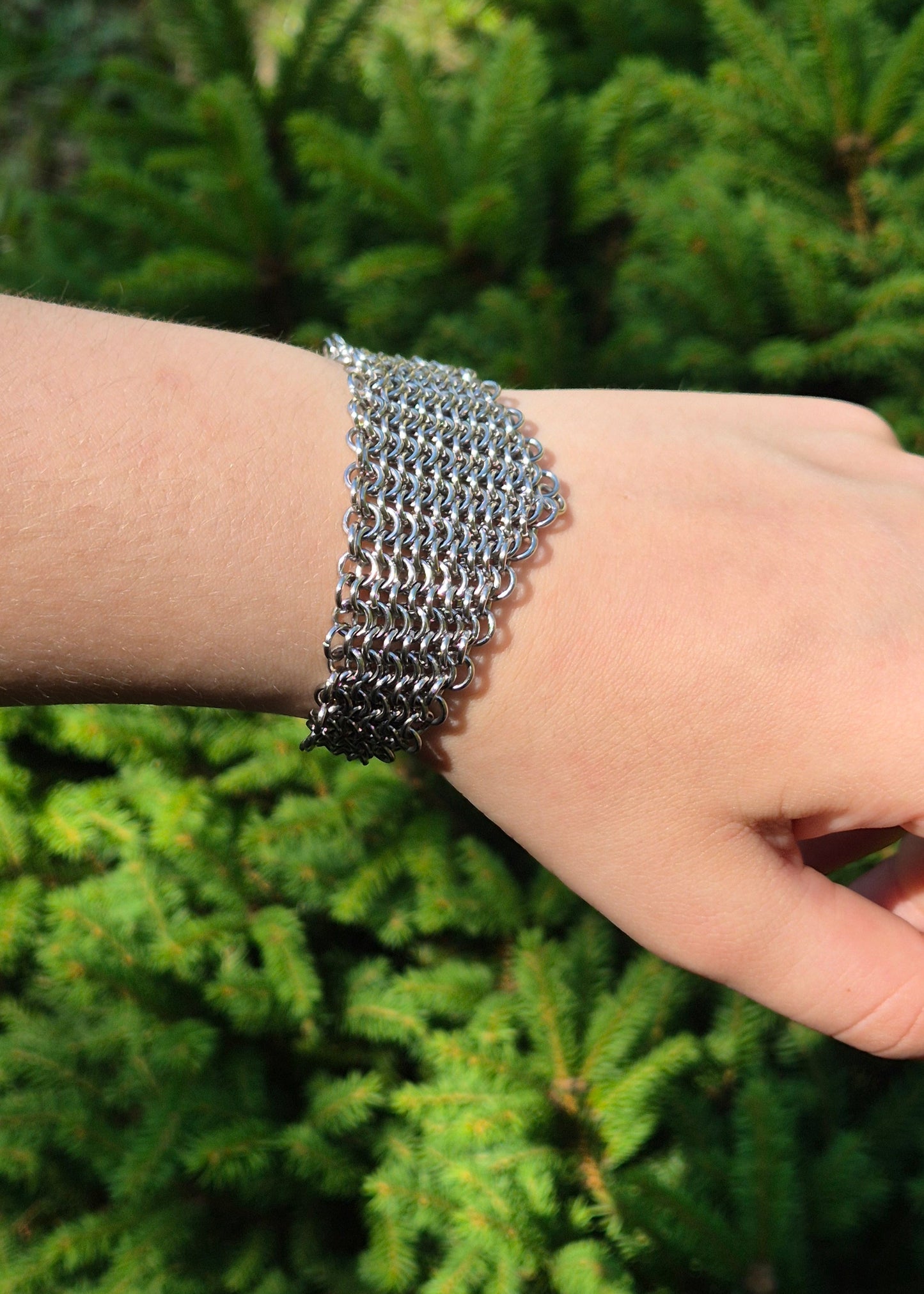 Zdjęcie nadgarstka z szeroką, ręcznie plecioną bransoletą typu chainmail wykonaną ze stali nierdzewnej w splocie europejskim 4 w 1. Bransoleta ma surowy, industrialny charakter i nawiązuje do stylu gotyckiego, alternatywnego, fantasy i średniowiecznego. Tłem jest zielony świerk podkreślający kontrast stali i natury.