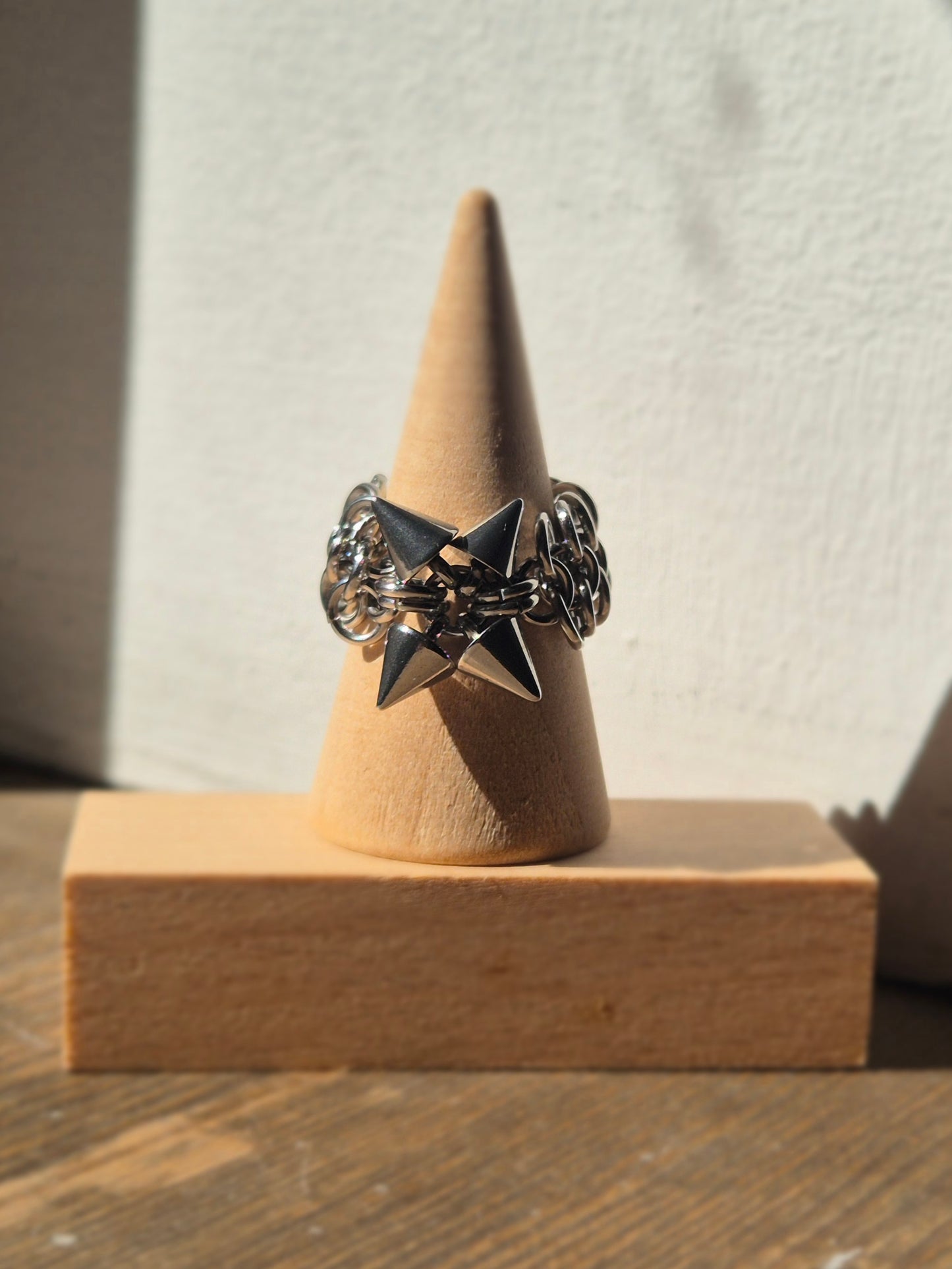 Zbliżenie na ręcznie wykonaną stalową obrączką w stylu chainmail, ozdobioną metalowymi ćwiekami w kształcie stożków. Pierścień spleciony jest w europejskim wzorze 4 w 1. Tłem jest biała ściana, która podkreśla surowy i alternatywny styl biżuterii.