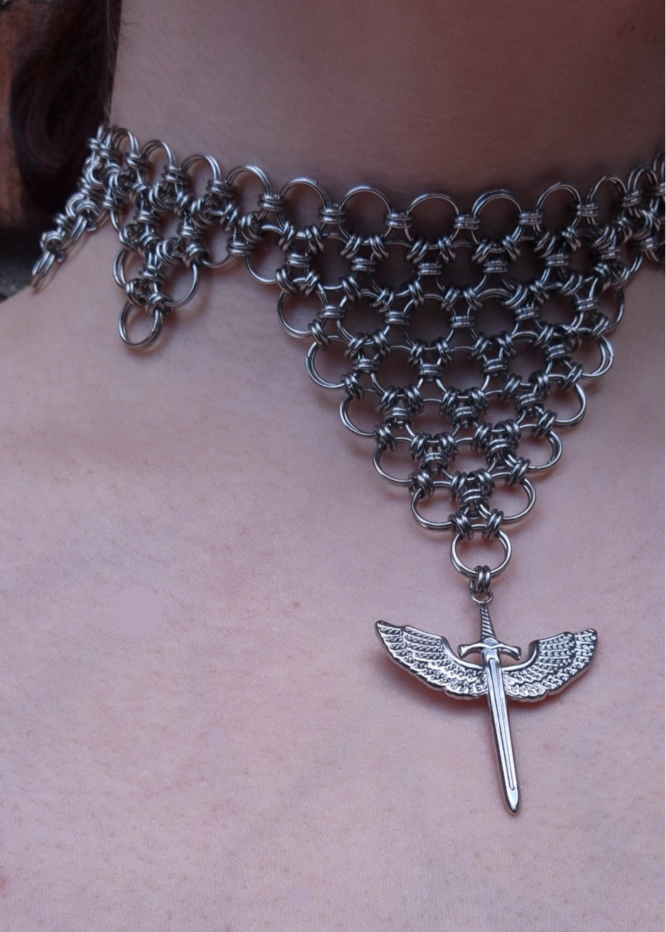 Zbliżenie na choker z kolczugi ze stali nierdzewnej w kształcie odwróconego trójkąta z zawieszką — skrzydlatym mieczem. Biżuteria handmade inspirowana estetyką wojowniczki i magii.