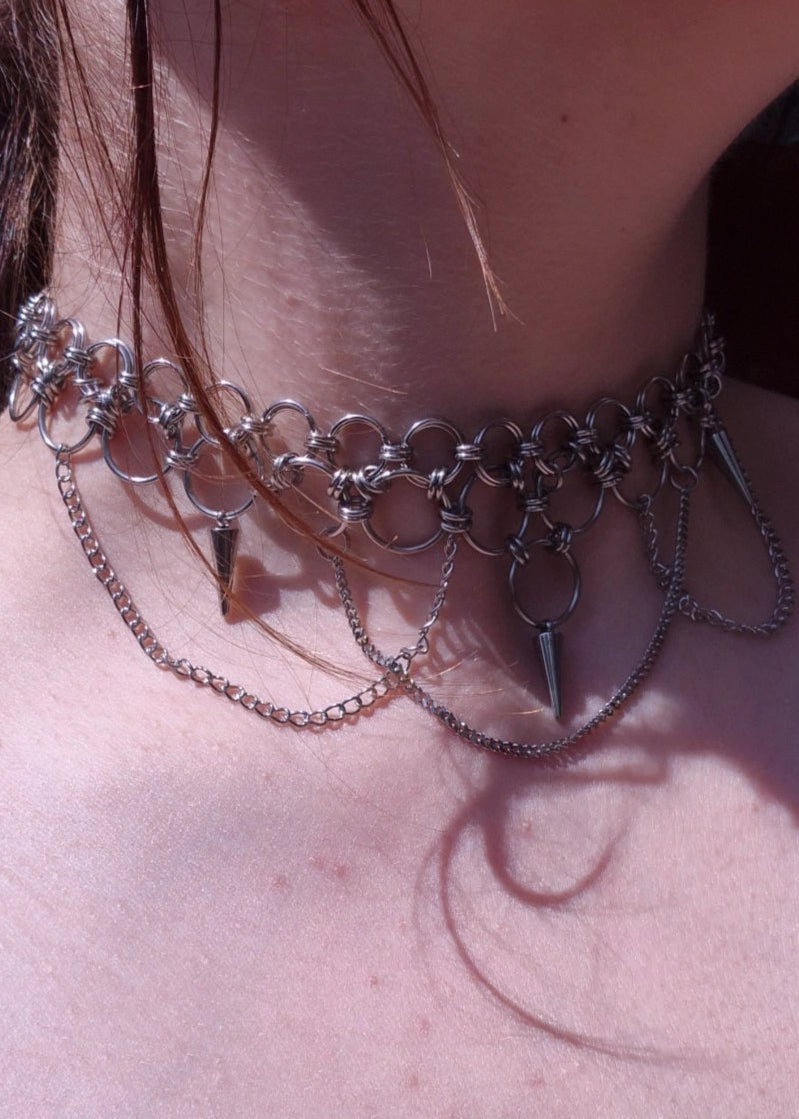 Zbliżenie na szyję z ręcznie robionym chokerem chainmaille ze stali nierdzewnej, ozdobionym kolcami, łańcuszkami i dużymi metalowymi ogniwami. Styl punk, industrial i alternatywny.