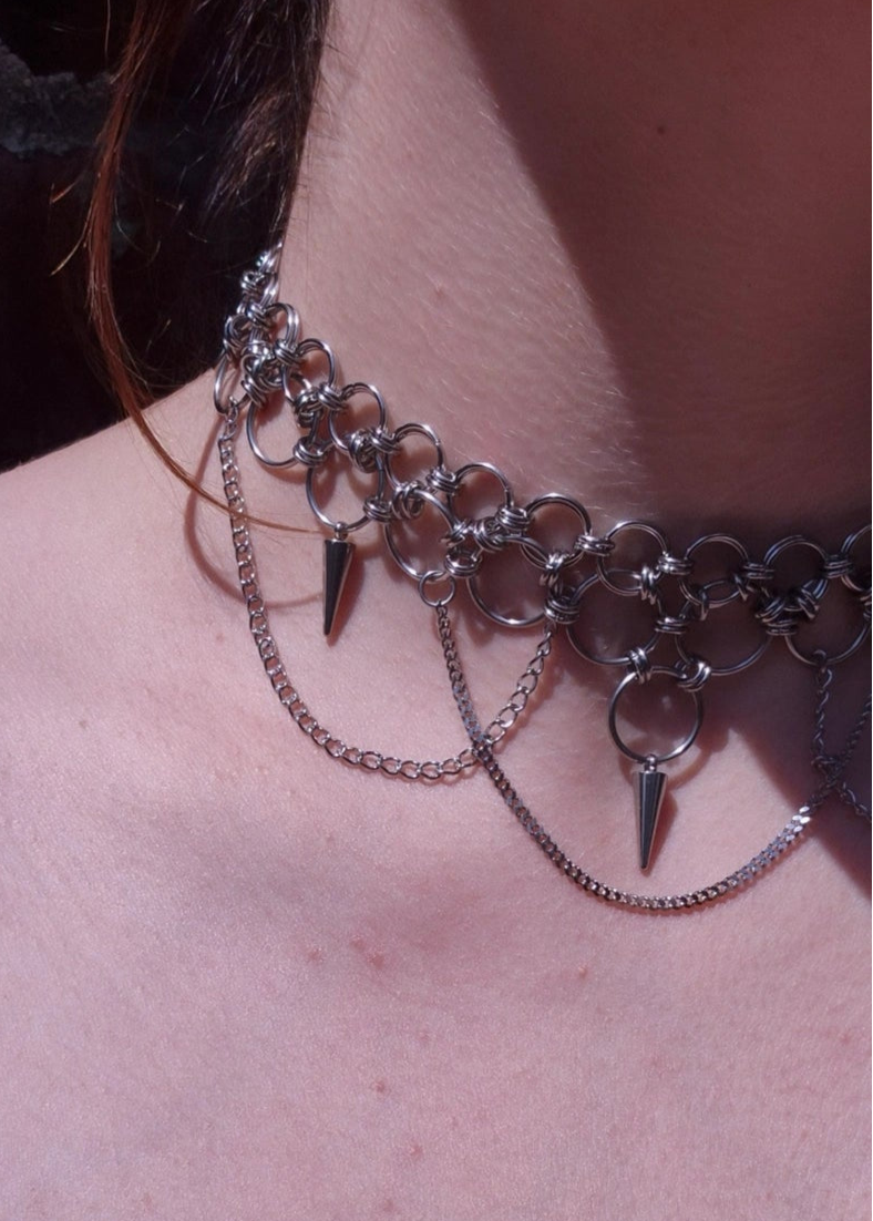 Zbliżenie na szyję z ręcznie robionym chokerem chainmaille ze stali nierdzewnej, ozdobionym kolcami, łańcuszkami i dużymi metalowymi ogniwami. Styl punk, industrial i alternatywny.
