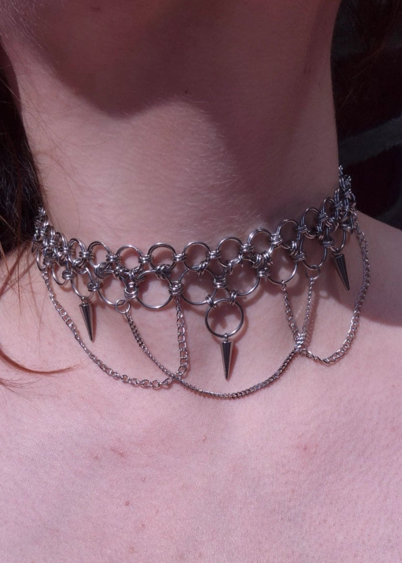 Zbliżenie na szyję z ręcznie robionym chokerem chainmaille ze stali nierdzewnej, ozdobionym kolcami, łańcuszkami i dużymi metalowymi ogniwami. Styl punk, industrial i alternatywny.