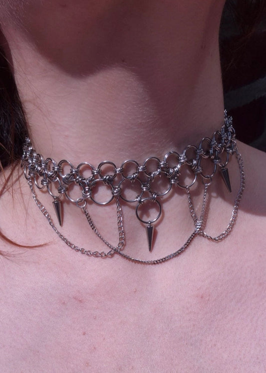 Zbliżenie na szyję z ręcznie robionym chokerem chainmaille ze stali nierdzewnej, ozdobionym kolcami, łańcuszkami i dużymi metalowymi ogniwami. Styl punk, industrial i alternatywny.