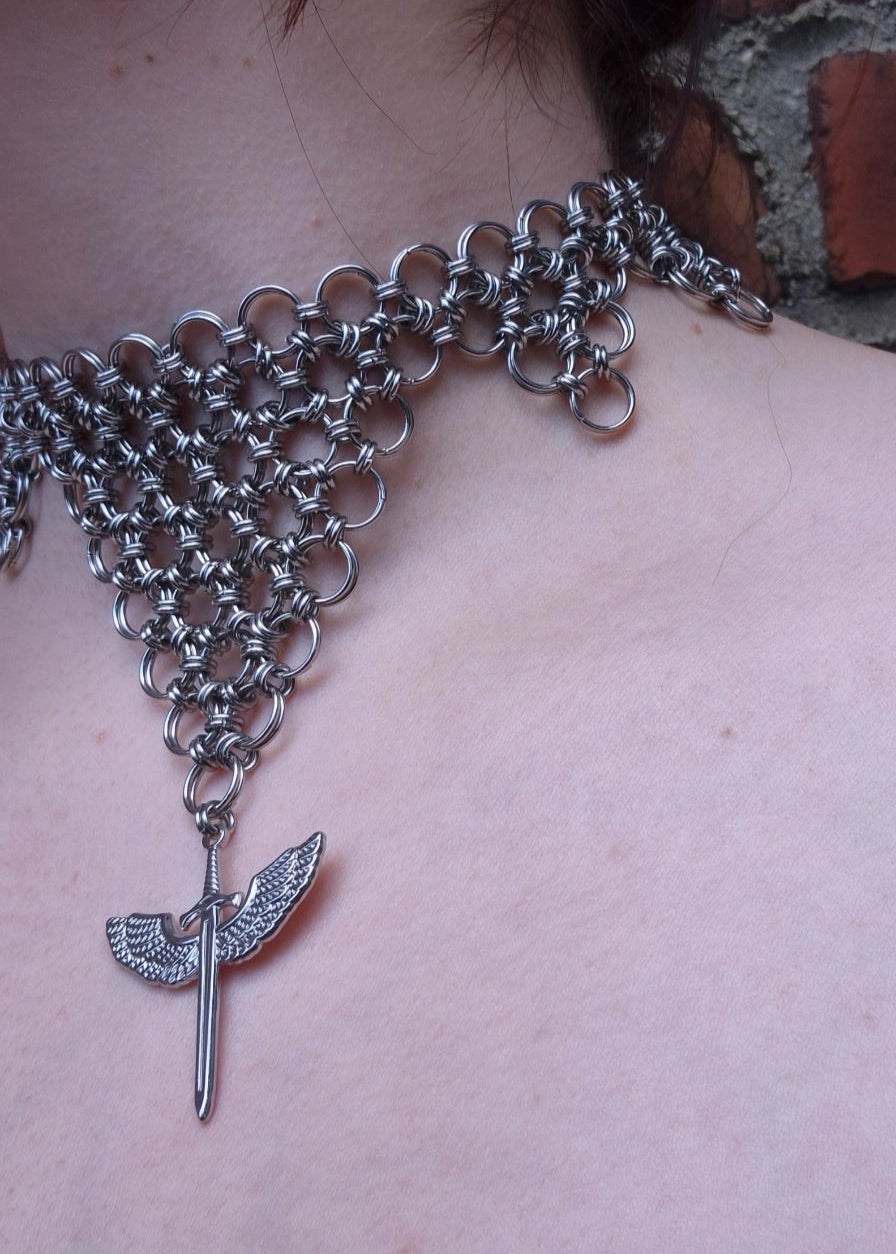 Zbliżenie na choker z kolczugi ze stali nierdzewnej w kształcie odwróconego trójkąta z zawieszką — skrzydlatym mieczem. Biżuteria handmade inspirowana estetyką wojowniczki i magii.