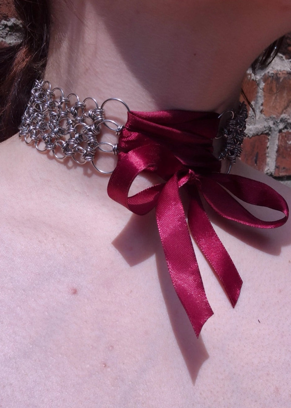 Zbliżenie na szyję z ręcznie robionym chokerem chainmaille ze stali nierdzewnej z wiązaniem z bordowej wstążki . Połączenie surowej kolczugi z delikatnością tkaniny, w klimacie dark romantic, lolita, alt fashion.