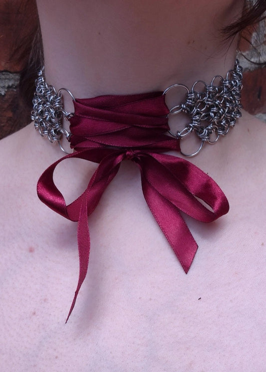Zbliżenie na szyję z ręcznie robionym chokerem chainmaille ze stali nierdzewnej z wiązaniem z bordowej wstążki . Połączenie surowej kolczugi z delikatnością tkaniny, w klimacie dark romantic, lolita, alt fashion.
