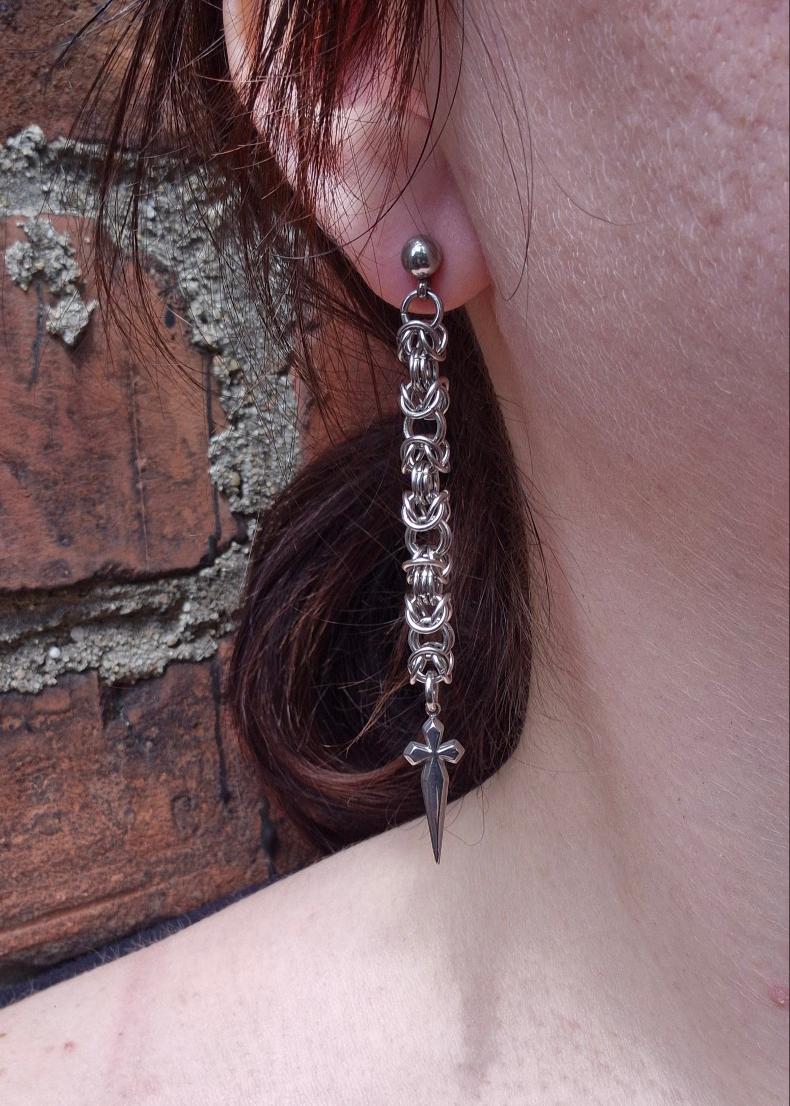 Zbliżenie na ucho osoby z kolczykiem wykonanym ze stalowych ogniw ułożonych w splot bizantyjski. Na końcu kolczyka zawieszony jest ciemny, gotycki krzyż. Tło to ceglana ściana z odpadającym tynkiem, dodająca industrialnego klimatu.