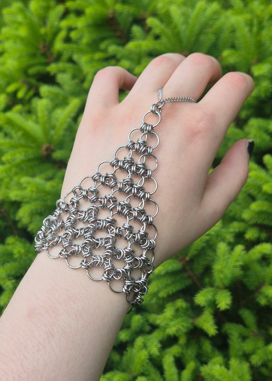 Zdjęcie przedstawia dłoń z założoną ręcznie plecioną bransoletką typu chainmail wykonaną ze stalowych ogniw. Biżuteria oplata nadgarstek i łączy się z palcem trójkątną siatką z kółeczek, tworząc lekki pancerz o gotyckim i alternatywnym charakterze. W tle intensywnie zielone igły świerku tworzą kontrast z chłodnym połyskiem stali.