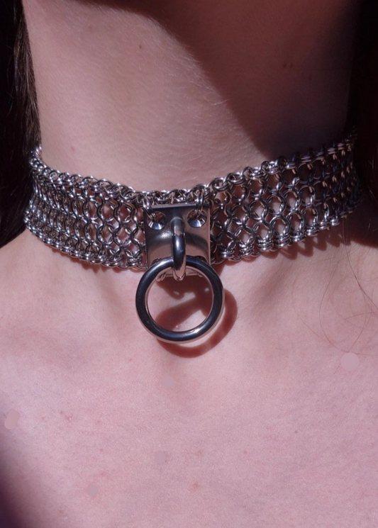 Zdjęcie przedstawia masywny choker w stylu chainmaille z ręcznie plecionych ogniwek ze stali nierdzewnej, noszony na szyi osoby o jasnej karnacji. W centrum chokera znajduje się duży stalowy O-ring zamocowany na solidnym, płaskim panelu. Design inspirowany stylem industrialnym oraz mroczną estetyką gotycką i alternatywną. Biżuteria przyciąga wzrok błyszczącą powierzchnią metalu i surowym charakterem.