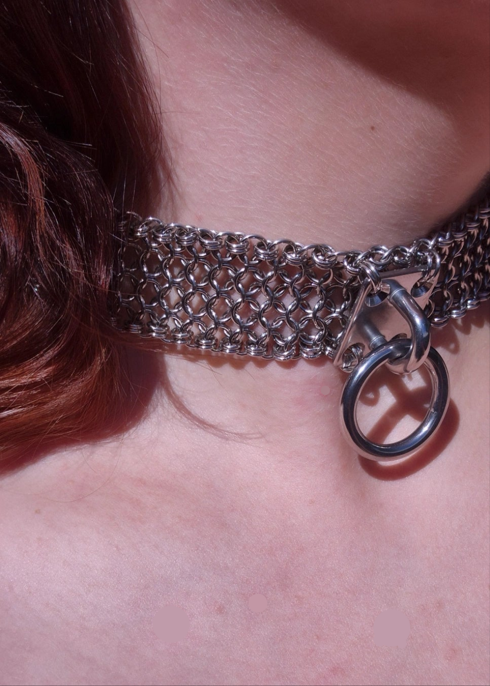 Zdjęcie przedstawia masywny choker w stylu chainmaille z ręcznie plecionych ogniwek ze stali nierdzewnej, noszony na szyi osoby o jasnej karnacji pod innym kątem niż poprzednie zdjęcie.  W centrum chokera znajduje się duży stalowy O-ring zamocowany na solidnym, płaskim panelu. Design inspirowany stylem industrialnym oraz mroczną estetyką gotycką i alternatywną. Biżuteria przyciąga wzrok błyszczącą powierzchnią metalu i surowym charakterem.