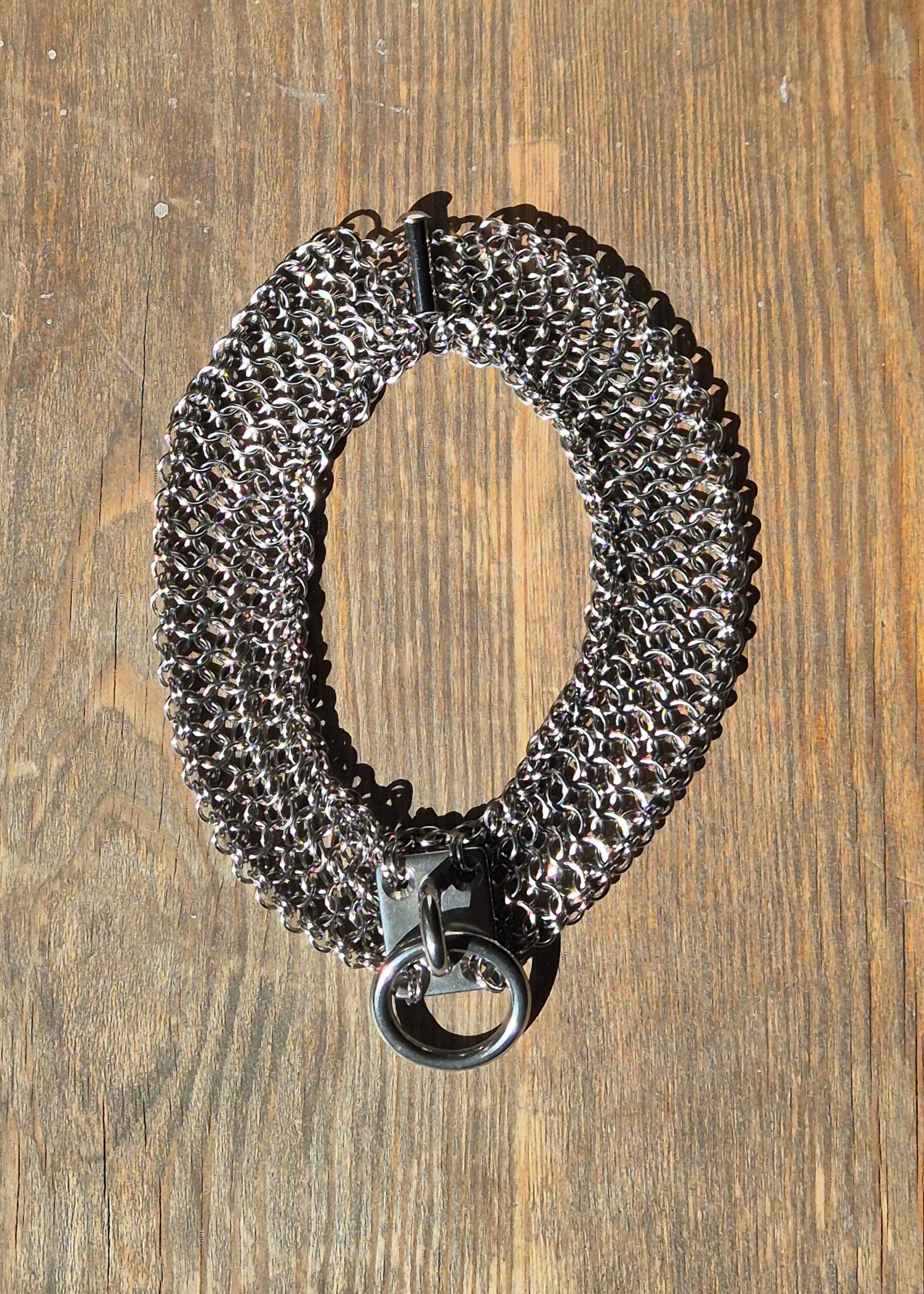 Zdjęcie przedstawia masywny choker w stylu chainmaille z ręcznie plecionych ogniwek ze stali nierdzewnej,  położony na drewnianej powierzchni. W centrum chokera znajduje się duży stalowy O-ring zamocowany na solidnym, płaskim panelu. Design inspirowany stylem industrialnym oraz mroczną estetyką gotycką i alternatywną. Biżuteria przyciąga wzrok błyszczącą powierzchnią metalu i surowym charakterem.