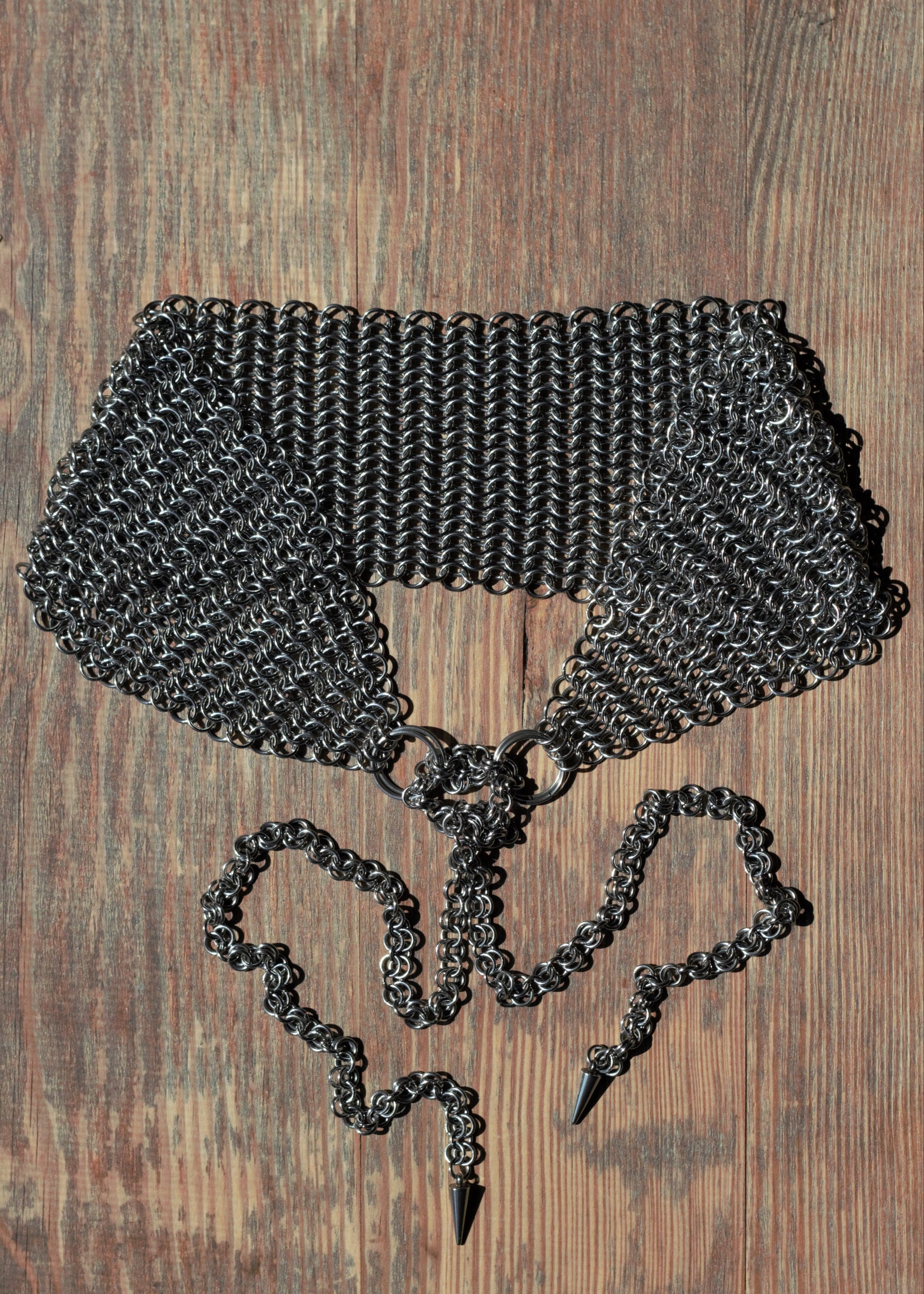 Szeroka opaska na głowę chainmail ze stali nierdzewnej ułożona na drewnianym podłożu, na końcach opaski znajdują się łańcuszki zakończone kolcami.