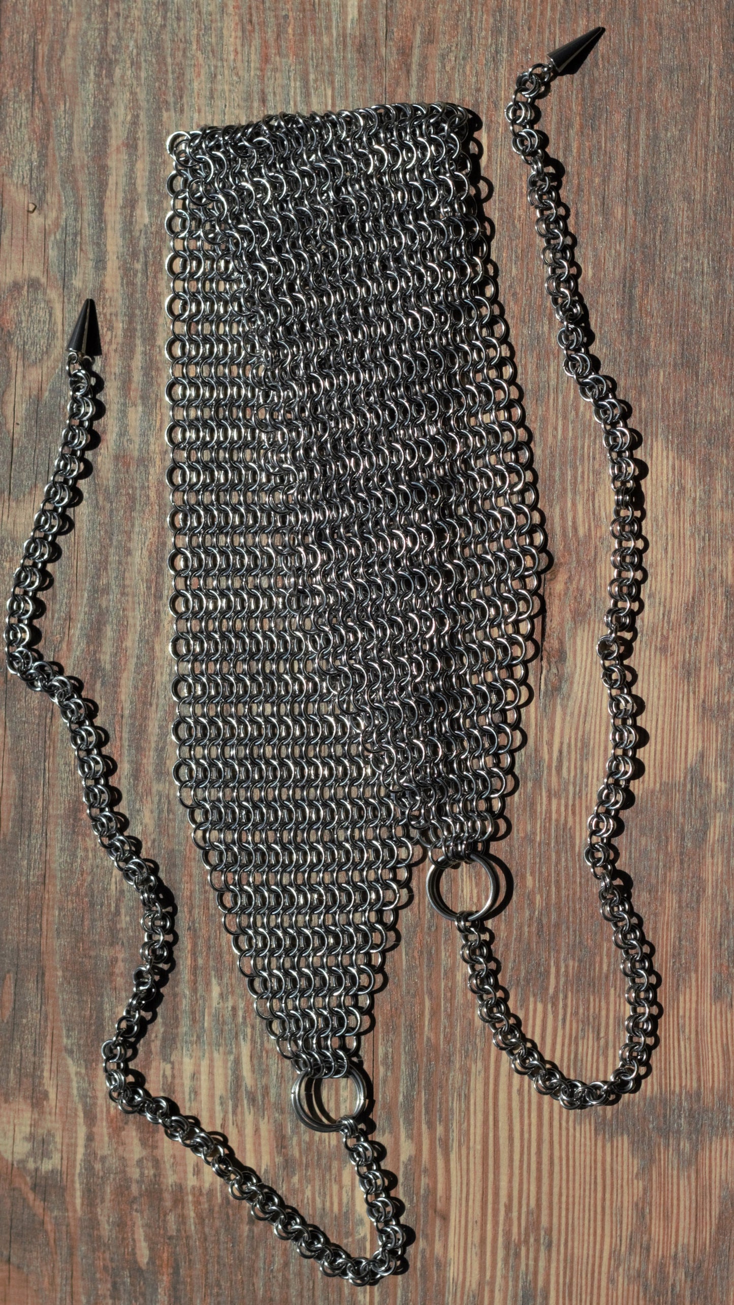 Szeroka opaska na głowę chainmail ze stali nierdzewnej ułożona na drewnianym podłożu, na końcach opaski znajdują się łańcuszki zakończone kolcami.