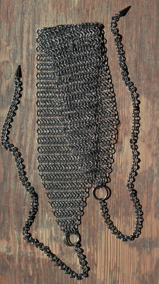 Szeroka opaska na głowę chainmail ze stali nierdzewnej ułożona na drewnianym podłożu, na końcach opaski znajdują się łańcuszki zakończone kolcami.