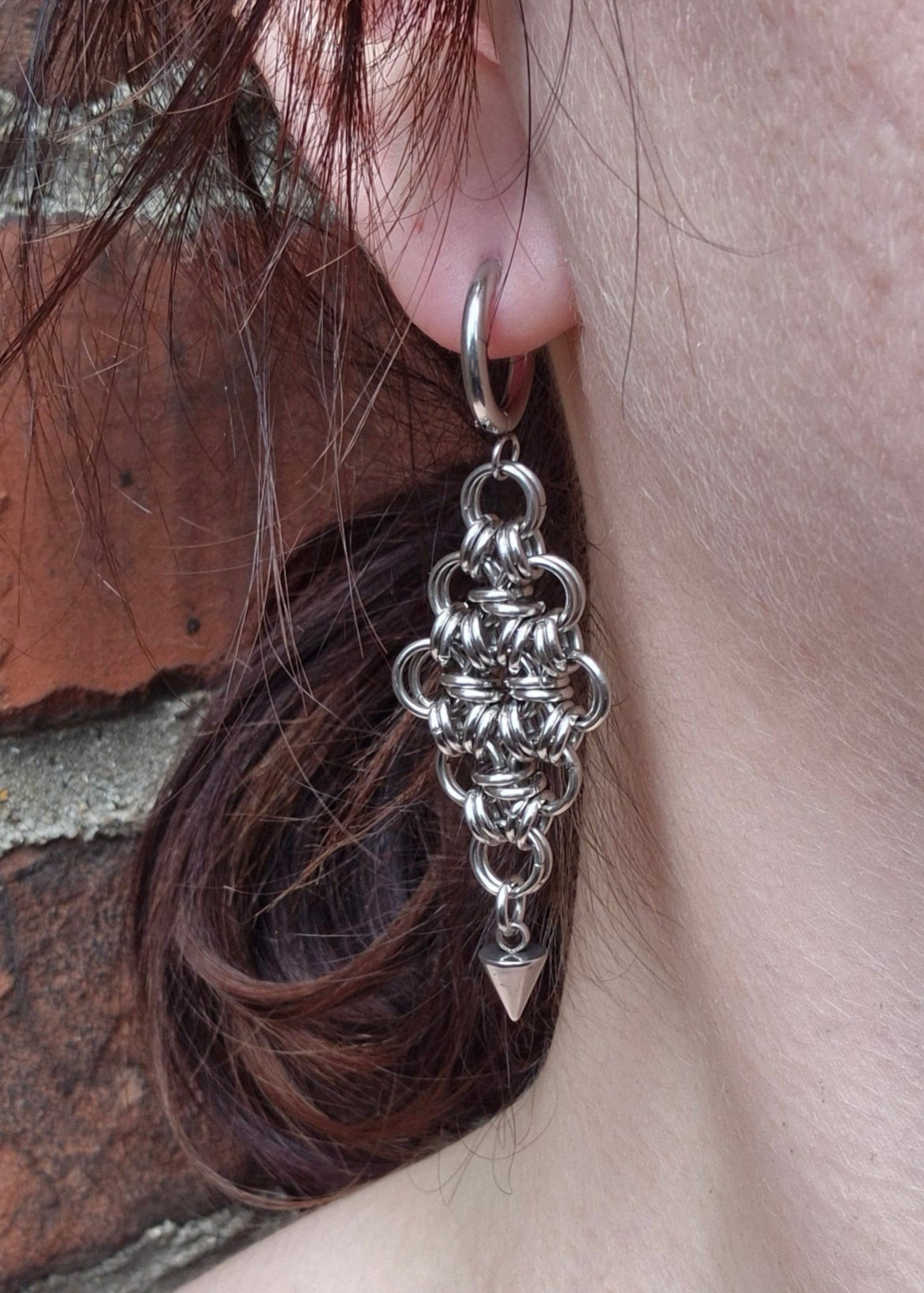 Zbliżenie na ucho z ręcznie robionym kolczykiem chainmaille ze stali nierdzewnej, o kształcie rombu z geometrycznym kolcem na dole. Biżuteria industrialna, handmade, alternatywna.