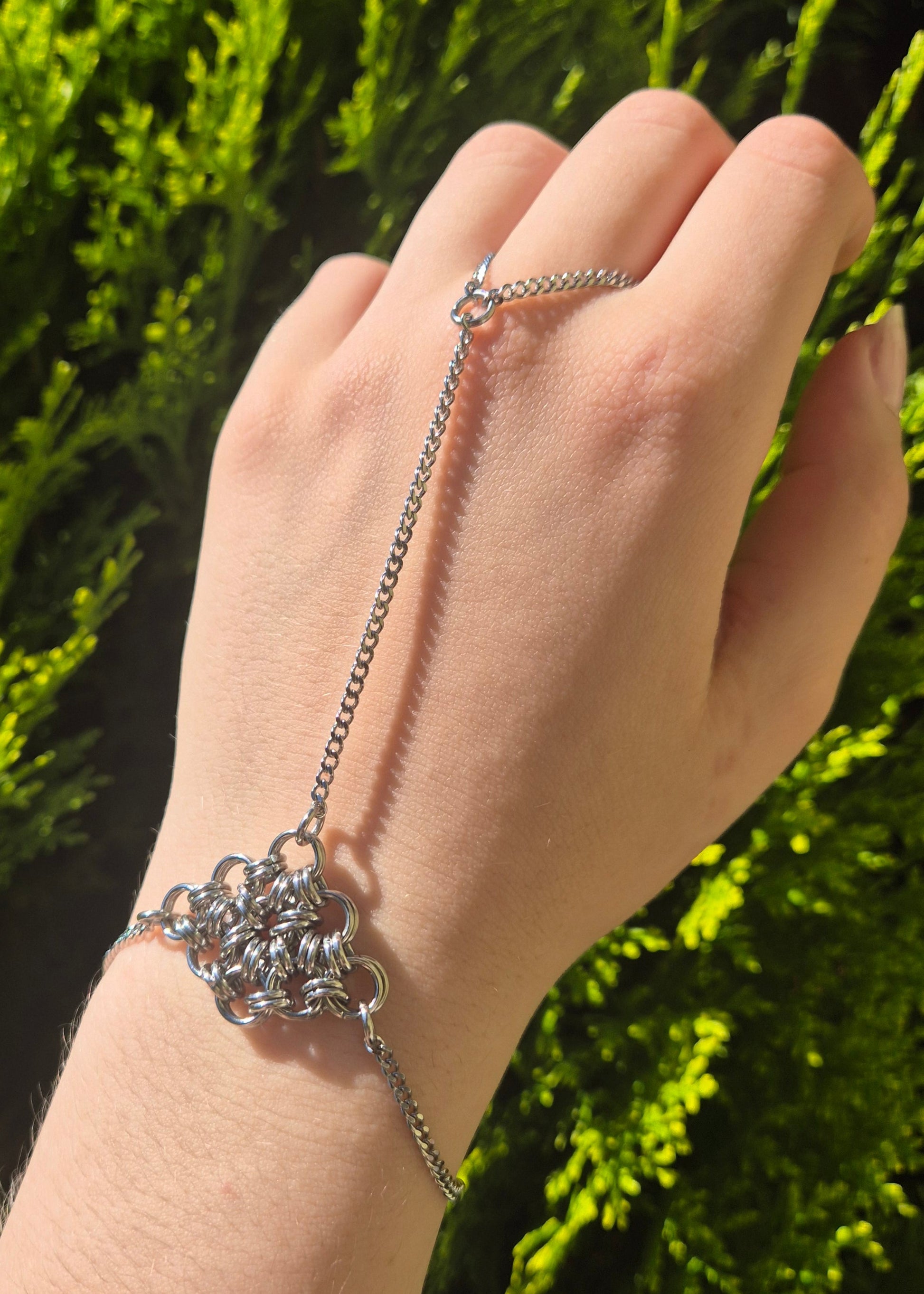 Srebrna bransoletka łącząca nadgarstek i palec, z ozdobnym elementem chainmail na grzbiecie dłoni, wykonana ze stali nierdzewnej, w słońcu na tle zielonych liści.