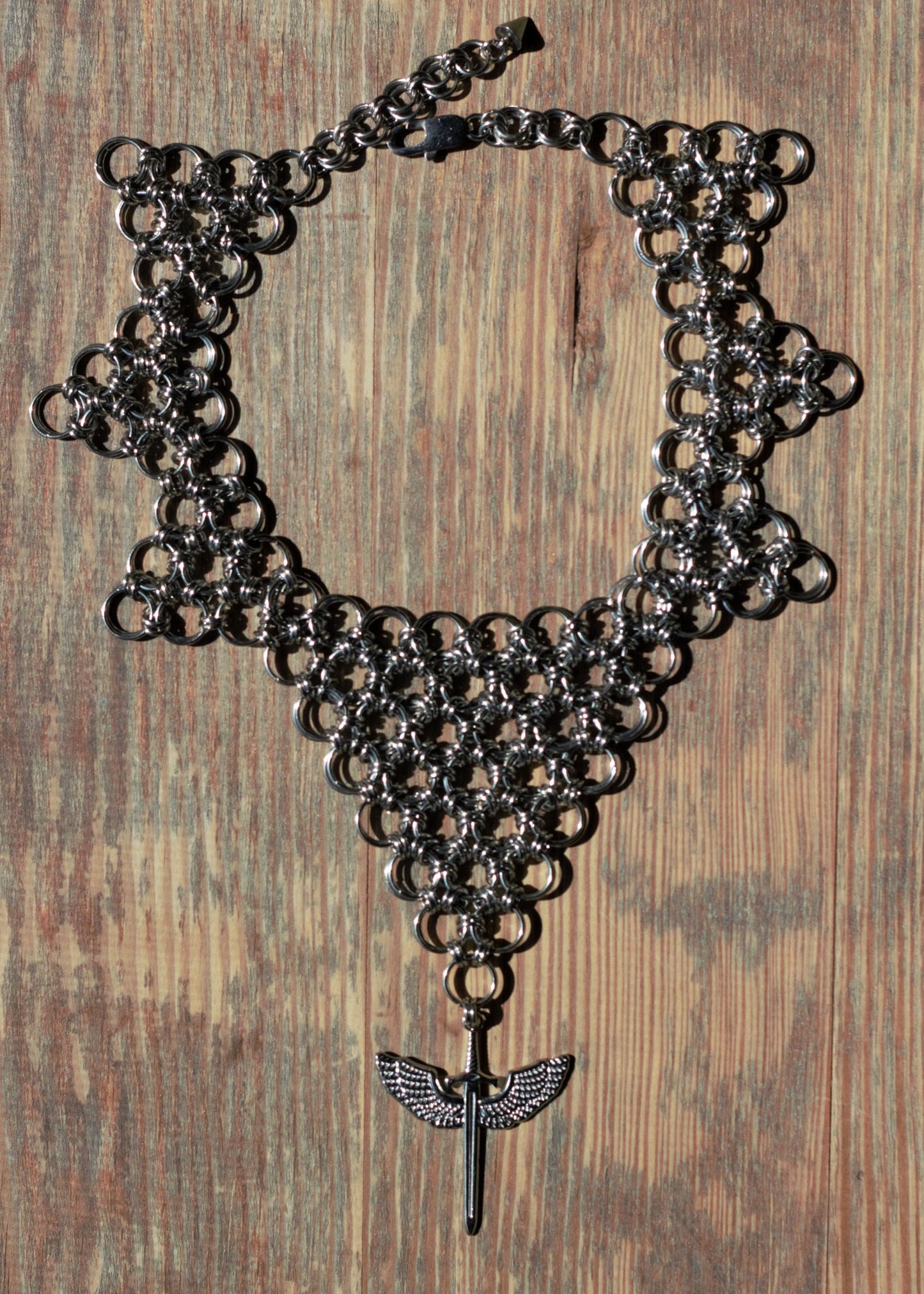 Choker ze stali nierdzewnej w kształcie odwróconego trójkąta z zawieszką - skrzydlatym mieczem, położony na drewnianej powierzchni.