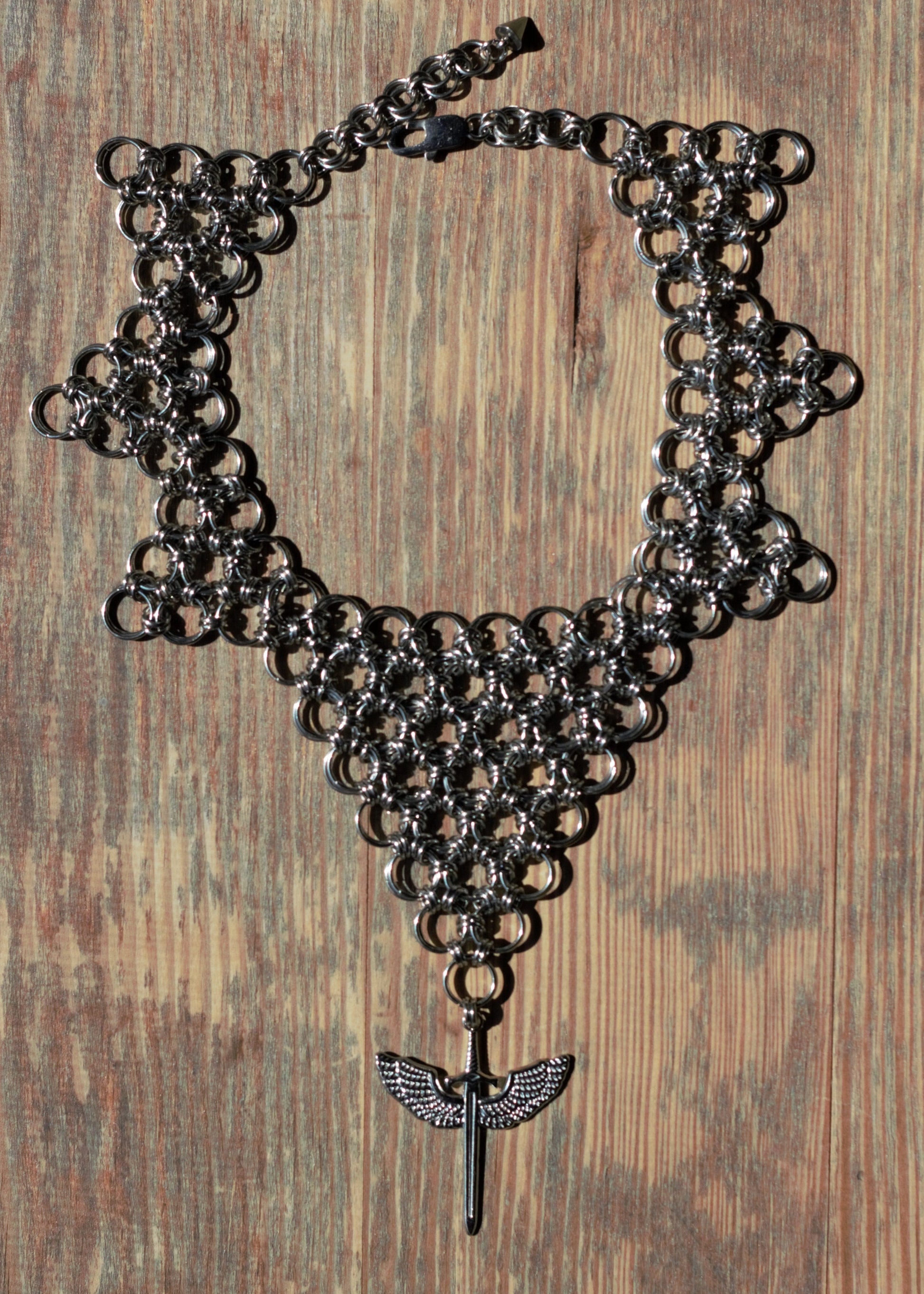 Choker ze stali nierdzewnej w kształcie odwróconego trójkąta z zawieszką - skrzydlatym mieczem, położony na drewnianej powierzchni.