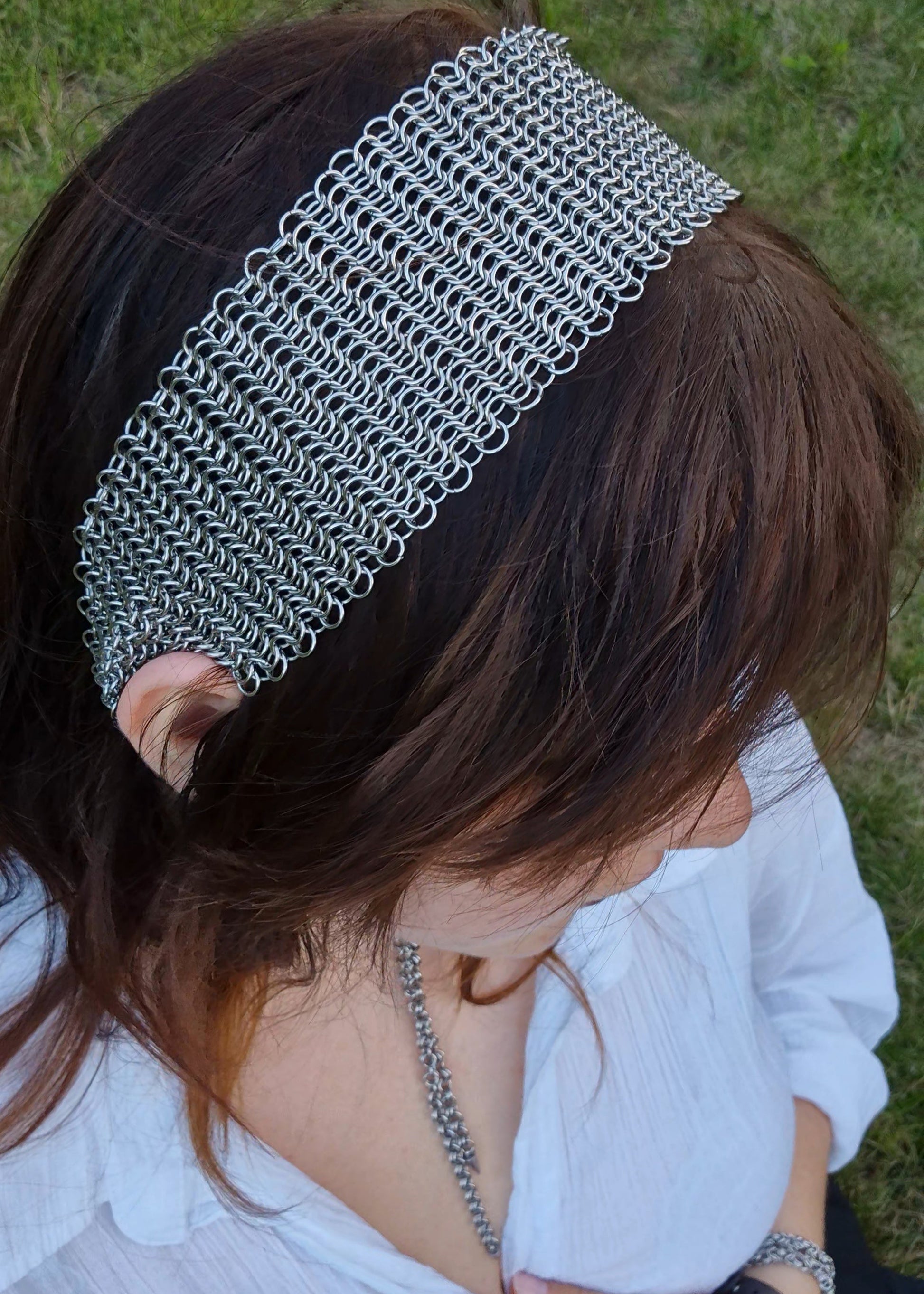 Zbliżenie na głowę kobiety z założoną szeroką opaską chainmaille wykonaną ręcznie ze stali nierdzewnej. Biżuteria alternatywna w stylu średniowiecznym i fantasy. W tle zielona trawa, stylizacja z białą koszulą