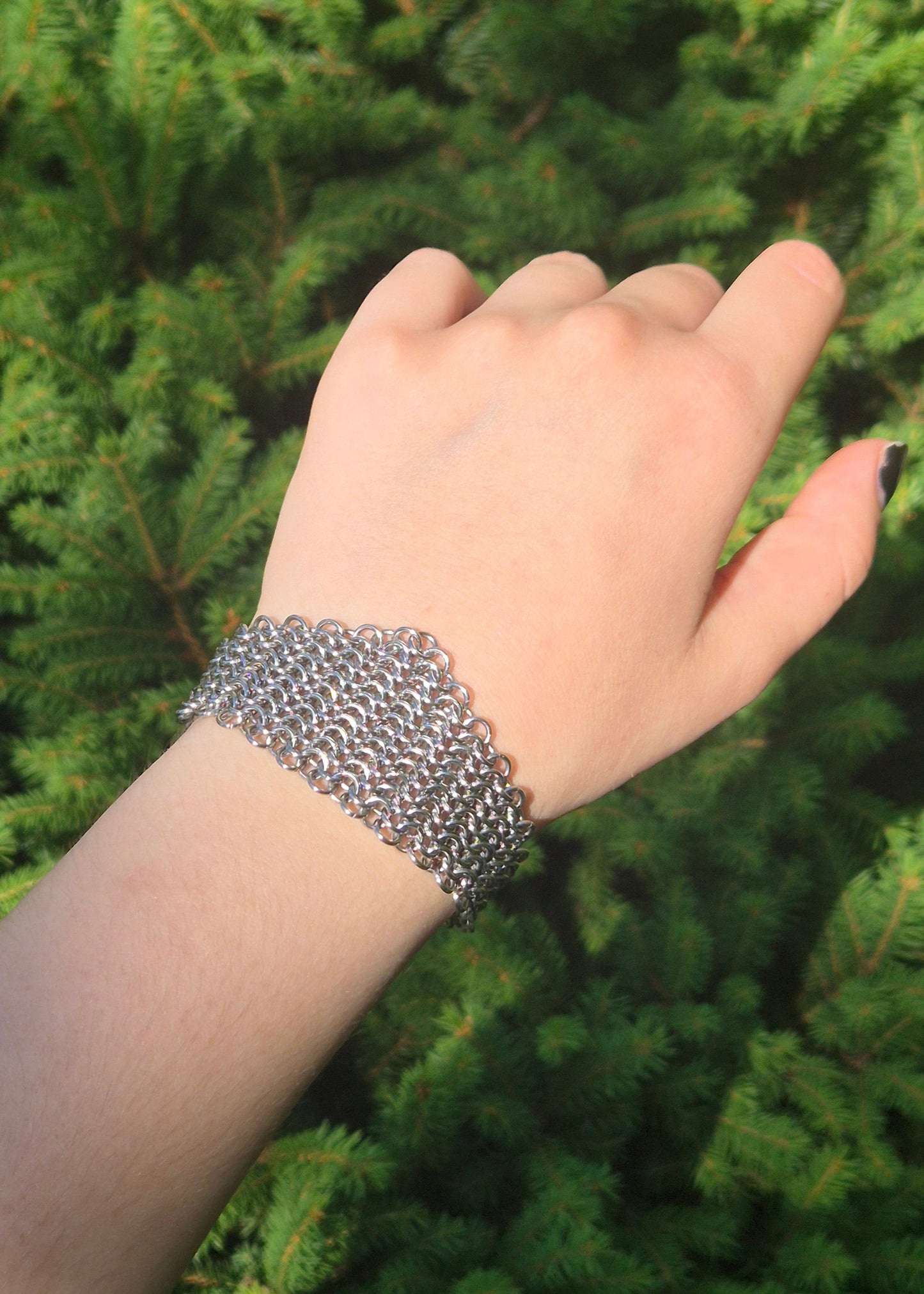 Zdjęcie nadgarstka z szeroką, ręcznie plecioną bransoletą typu chainmail wykonaną ze stali nierdzewnej w splocie europejskim 4 w 1. Bransoleta ma surowy, industrialny charakter i nawiązuje do stylu gotyckiego, alternatywnego, fantasy i średniowiecznego. Tłem jest zielony świerk podkreślający kontrast stali i natury.