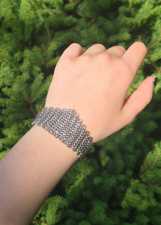 Zdjęcie nadgarstka z szeroką, ręcznie plecioną bransoletą typu chainmail wykonaną ze stali nierdzewnej w splocie europejskim 4 w 1. Bransoleta ma surowy, industrialny charakter i nawiązuje do stylu gotyckiego, alternatywnego, fantasy i średniowiecznego. Tłem jest zielony świerk podkreślający kontrast stali i natury.