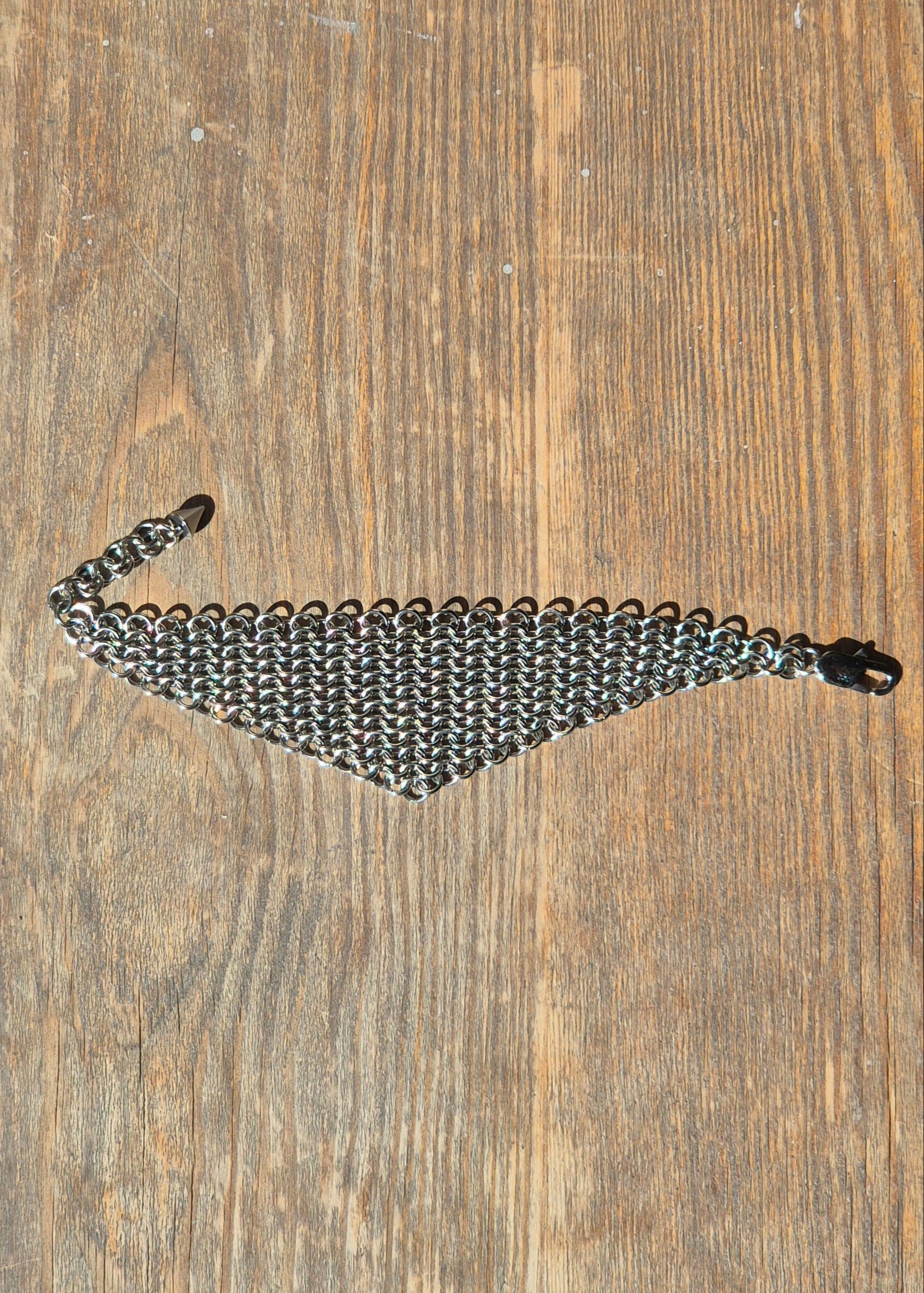 Zdjęcie szerokiej, ręcznie plecionej bransolet typu chainmail wykonana ze stali nierdzewnej w splocie europejskim 4 w 1. Bransoleta ma surowy, industrialny charakter i nawiązuje do stylu gotyckiego, alternatywnego, fantasy i średniowiecznego. Tłem jest drewniana powierzchnia