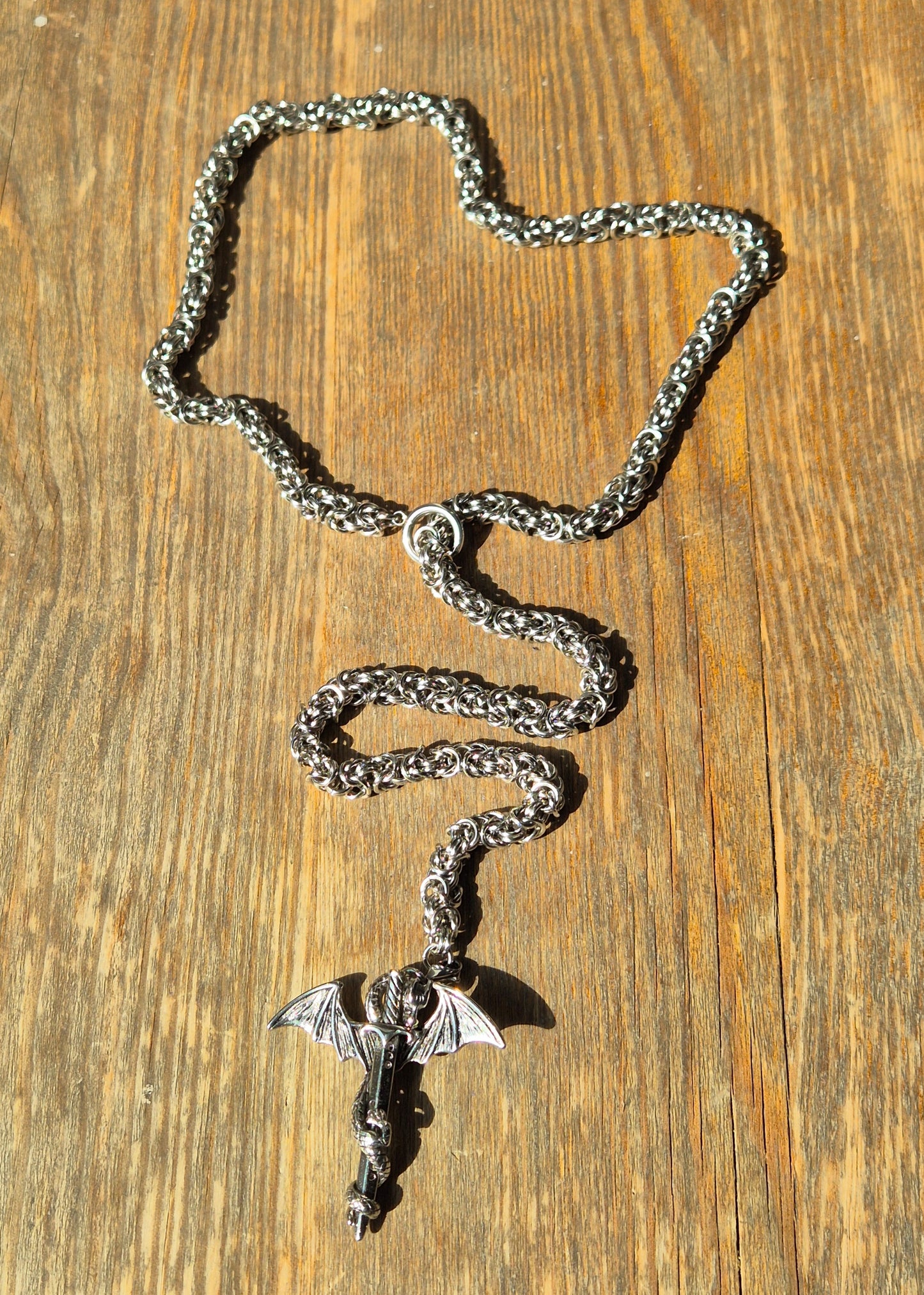 Naszyjnik z wisiorkiem smoka oplecionego wokół miecza, położony na rustykalnym drewnie, w mocnym świetle dziennym.