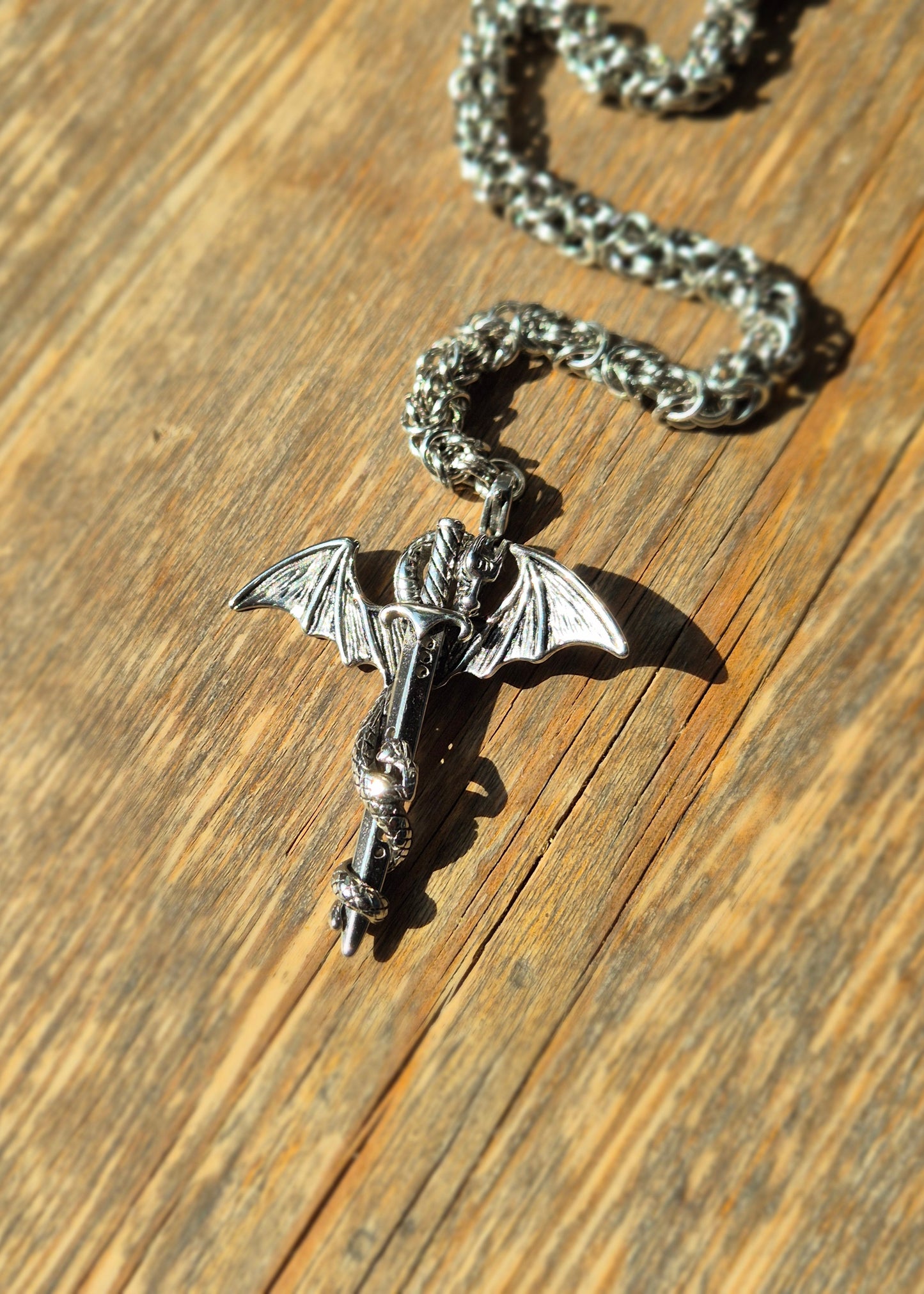 Naszyjnik z wisiorkiem smoka oplecionego wokół miecza, położony na rustykalnym drewnie, w mocnym świetle dziennym.