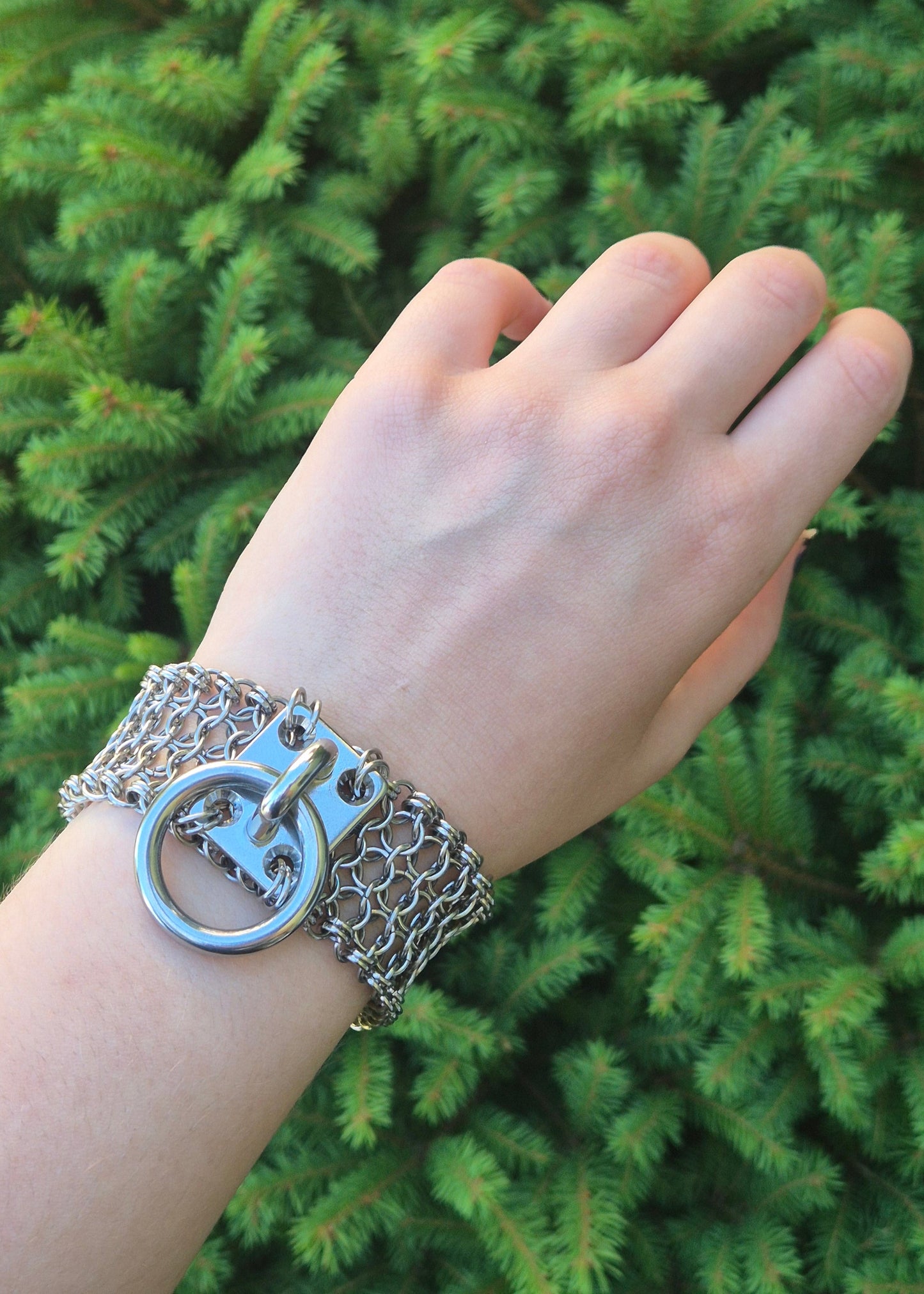 Zdjęcie przedstawia masywną bransoletę w stylu chainmaille z ręcznie plecionych ogniwek ze stali nierdzewnej, założona na nadgarsku osoby o jasnej karnacji, w tle zielone krzaki. Na środku znajduje się duży stalowy O-ring zamocowany na solidnym, płaskim panelu. Design inspirowany stylem industrialnym oraz mroczną estetyką gotycką i alternatywną. Biżuteria przyciąga wzrok błyszczącą powierzchnią metalu i surowym charakterem.