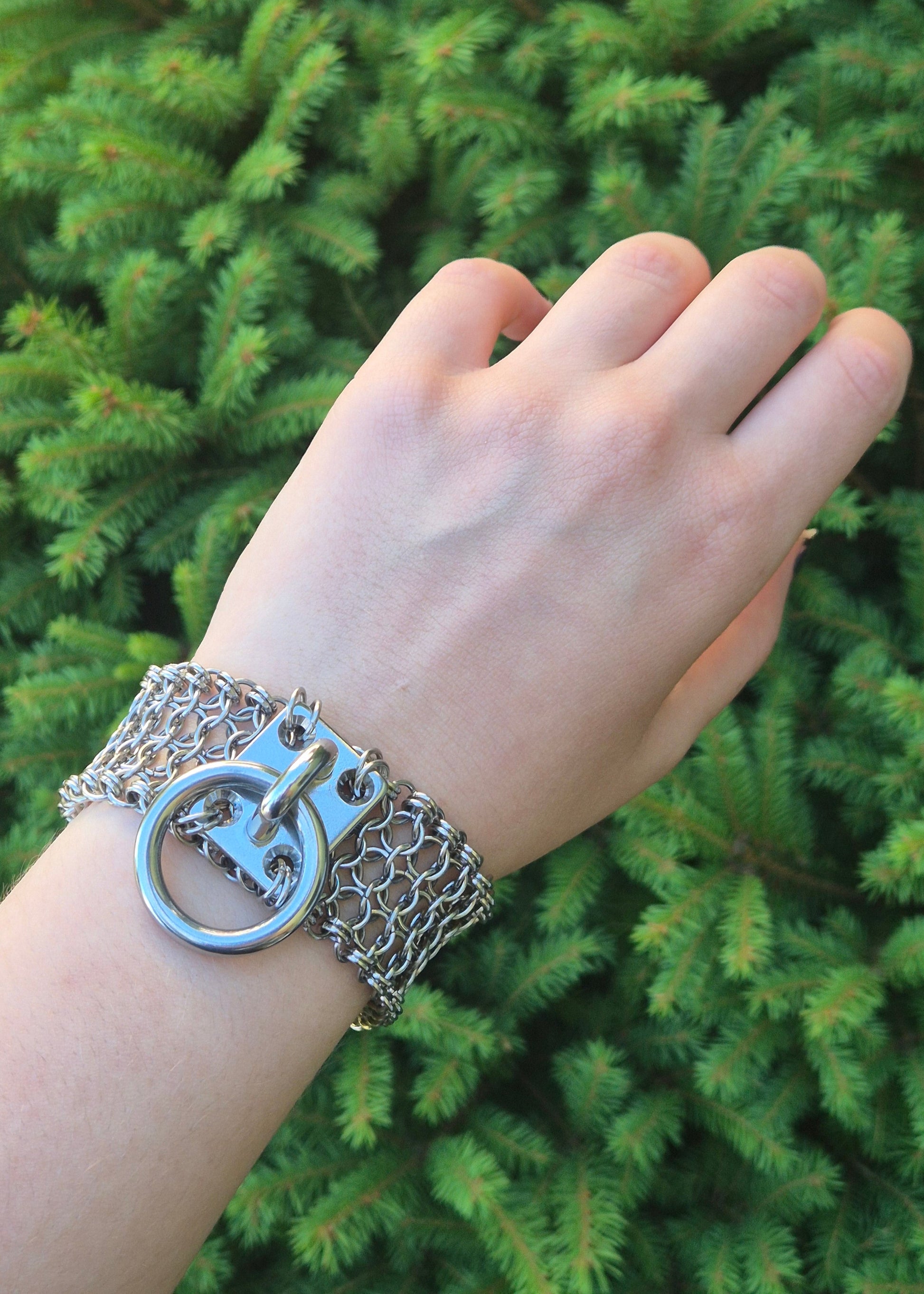 Zdjęcie przedstawia masywną bransoletę w stylu chainmaille z ręcznie plecionych ogniwek ze stali nierdzewnej, założona na nadgarsku osoby o jasnej karnacji, w tle zielone krzaki. Na środku znajduje się duży stalowy O-ring zamocowany na solidnym, płaskim panelu. Design inspirowany stylem industrialnym oraz mroczną estetyką gotycką i alternatywną. Biżuteria przyciąga wzrok błyszczącą powierzchnią metalu i surowym charakterem.