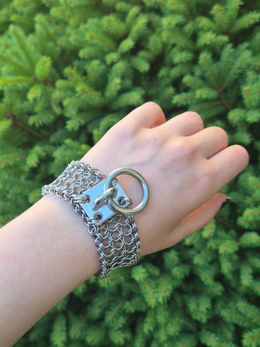 Zdjęcie przedstawia masywną bransoletę w stylu chainmaille z ręcznie plecionych ogniwek ze stali nierdzewnej, założona na nadgarsku osoby o jasnej karnacji, w tle zielone krzaki. Na środku znajduje się duży stalowy O-ring zamocowany na solidnym, płaskim panelu. Design inspirowany stylem industrialnym oraz mroczną estetyką gotycką i alternatywną. Biżuteria przyciąga wzrok błyszczącą powierzchnią metalu i surowym charakterem.