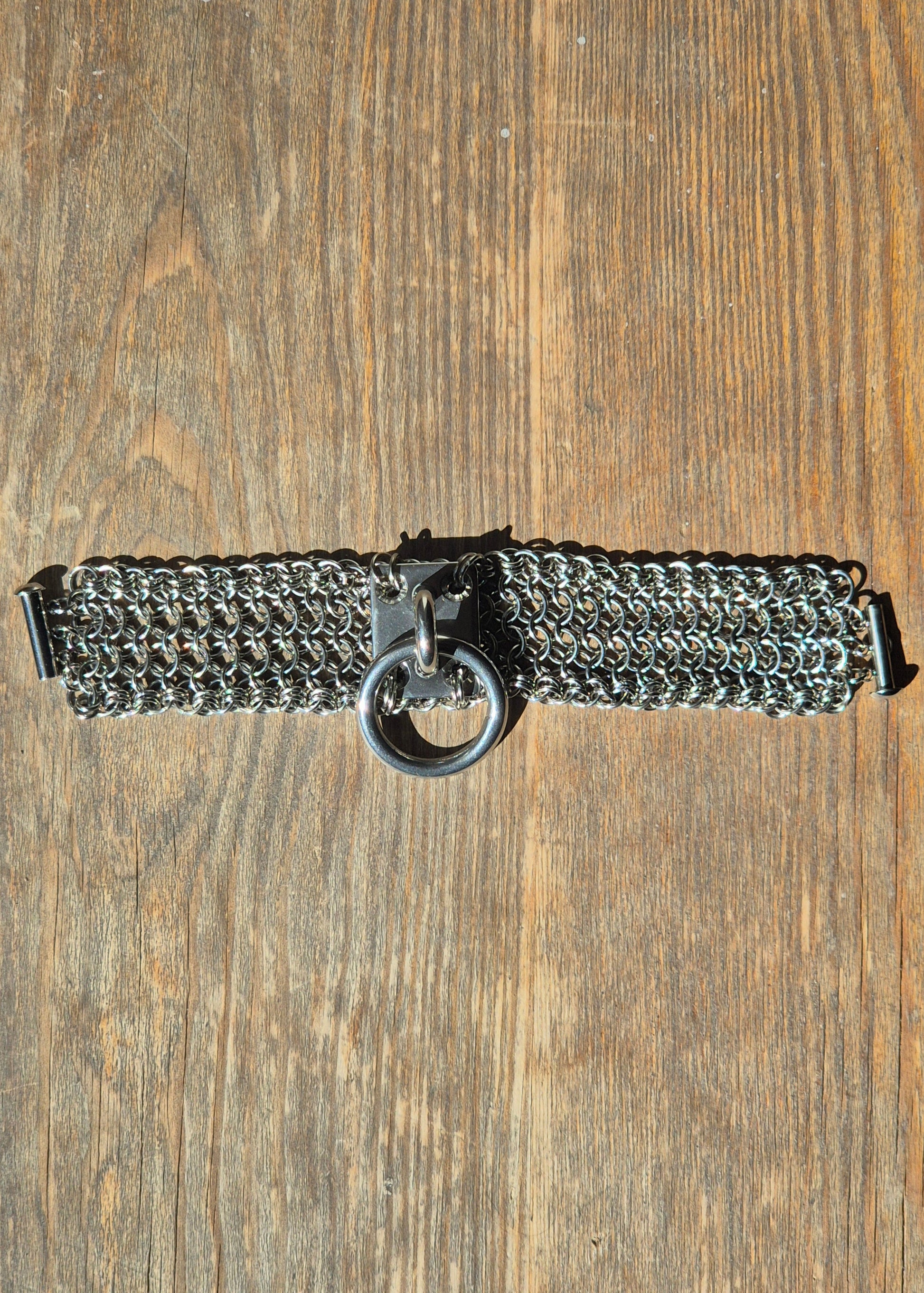 Zdjęcie przedstawia masywną bransoletę w stylu chainmaille z ręcznie plecionych ogniwek ze stali nierdzewnej, położona na drewnianej powierzchni. Na środku znajduje się duży stalowy O-ring zamocowany na solidnym, płaskim panelu. Design inspirowany stylem industrialnym oraz mroczną estetyką gotycką i alternatywną. Biżuteria przyciąga wzrok błyszczącą powierzchnią metalu i surowym charakterem.