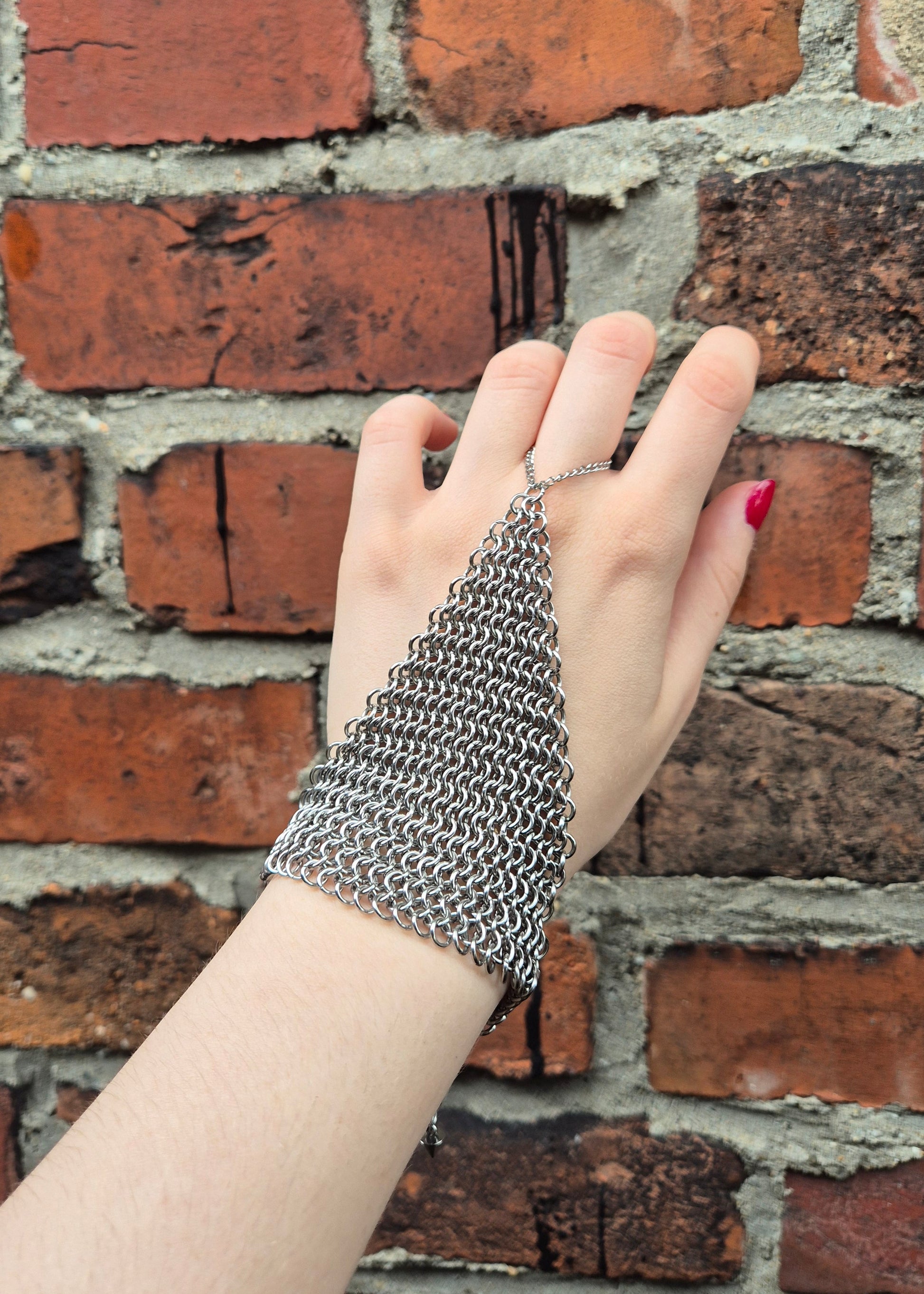 Dłoń ubrana w ręcznie wykonaną stalową rękawiczkę chainmail w splocie 4-w-1, z zapięciem na nadgarstek. Prezentowane na tle rustykalnej ściany z cegieł. Na kciuku osoby noszącej rękawice widoczny czerwony lakier do paznokci. Idealne dla rekonstrukcji historycznych, LARP, cosplayu lub stylizacji gotyckich/steampunkowych.