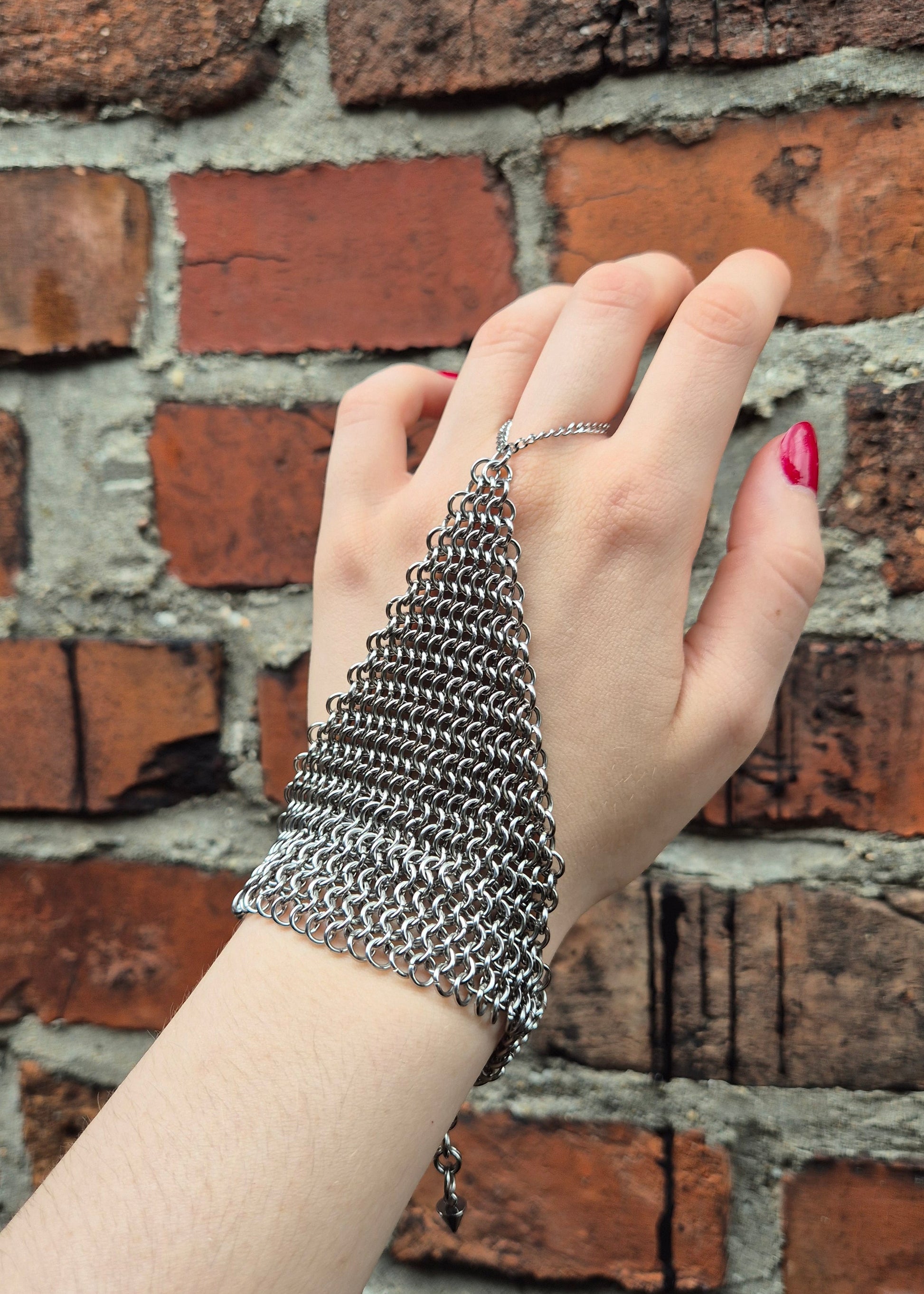 Dłoń ubrana w ręcznie wykonaną stalową rękawiczkę chainmail w splocie 4-w-1, z zapięciem na nadgarstek. Prezentowane na tle rustykalnej ściany z cegieł. Na kciuku osoby noszącej rękawice widoczny czerwony lakier do paznokci. Idealne dla rekonstrukcji historycznych, LARP, cosplayu lub stylizacji gotyckich/steampunkowych.