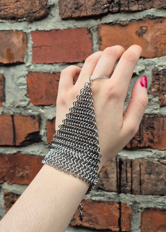 Dłoń ubrana w ręcznie wykonaną stalową rękawiczkę chainmail w splocie 4-w-1, z zapięciem na nadgarstek. Prezentowane na tle rustykalnej ściany z cegieł. Na kciuku osoby noszącej rękawice widoczny czerwony lakier do paznokci. Idealne dla rekonstrukcji historycznych, LARP, cosplayu lub stylizacji gotyckich/steampunkowych.