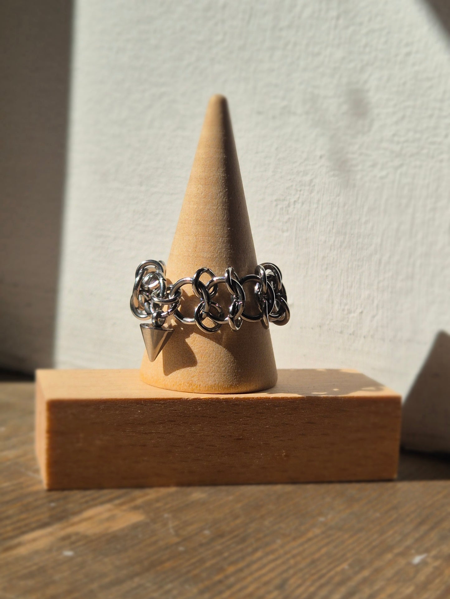 Zbliżenie na ręcznie wykonaną stalową obrączką w stylu chainmail, ozdobioną metalowym ćwiekiem w kształcie stożków. Pierścień założony na drewniany stożek. Tłem jest biała ściana, która podkreśla surowy i alternatywny styl biżuterii.