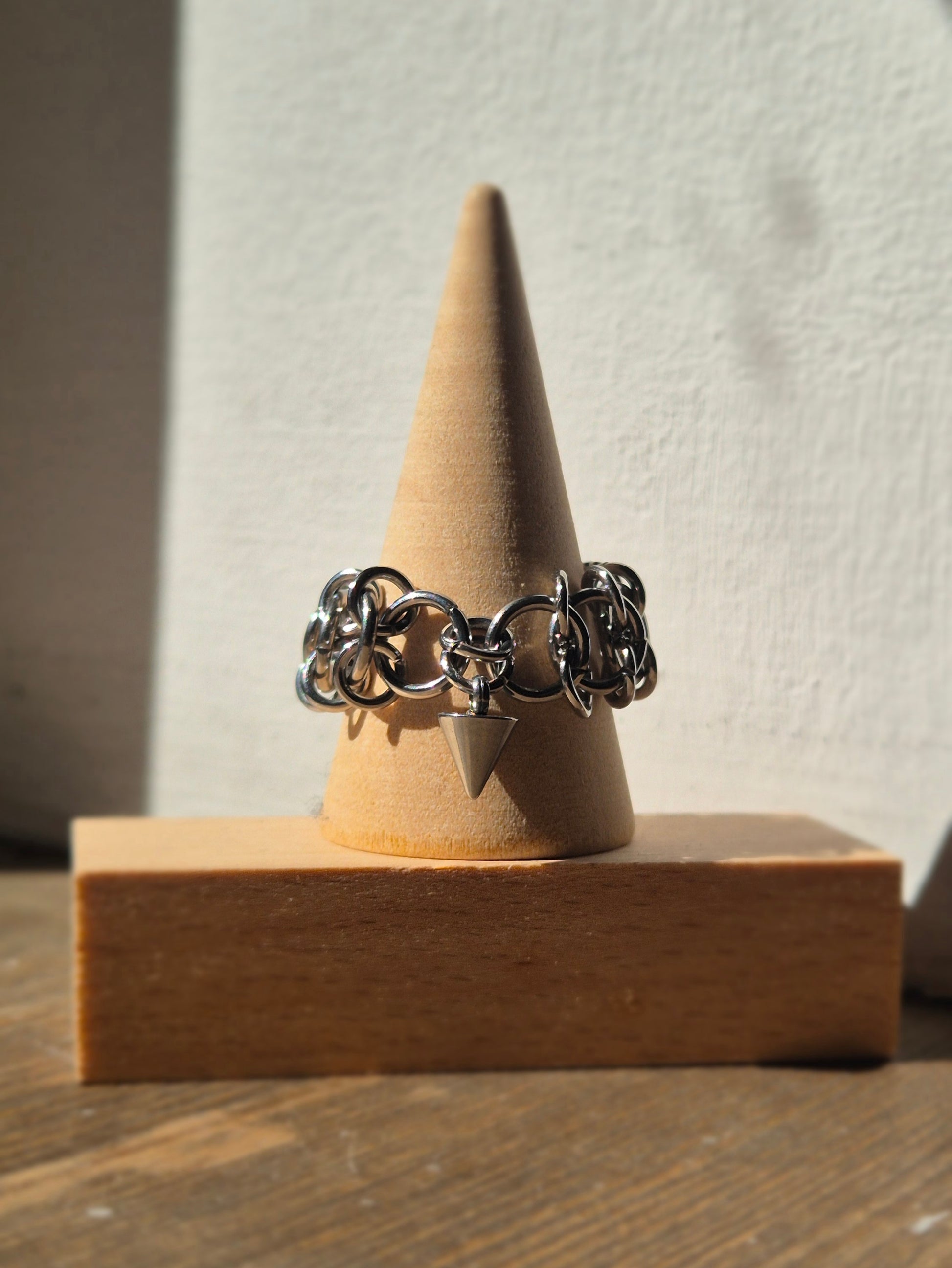 Zbliżenie na ręcznie wykonaną stalową obrączką w stylu chainmail, ozdobioną metalowym ćwiekiem w kształcie stożków. Pierścień założony na drewniany stożek. Tłem jest biała ściana, która podkreśla surowy i alternatywny styl biżuterii.