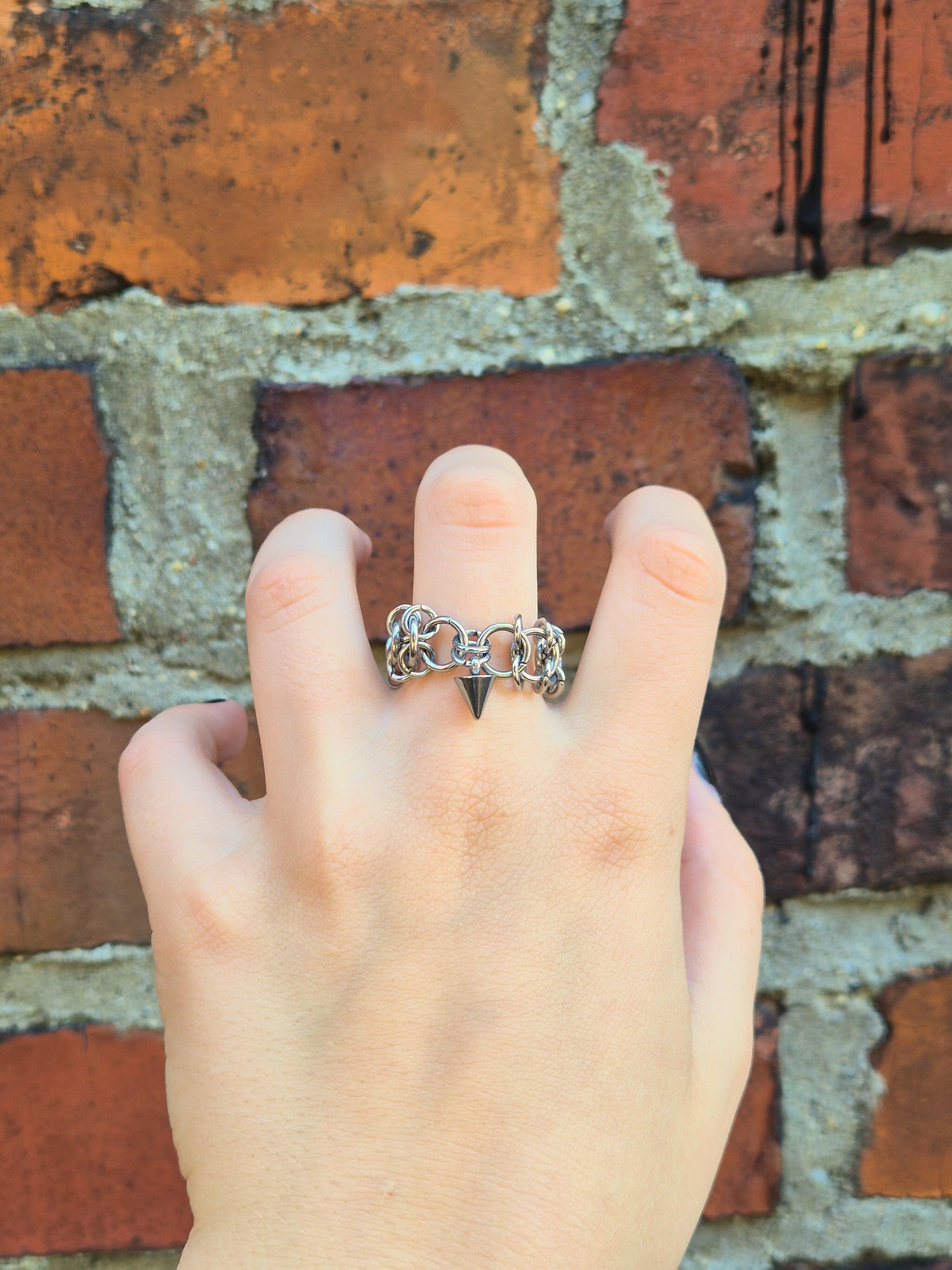 Zbliżenie na dłoń z ręcznie wykonaną stalową obrączką w stylu chainmail, ozdobioną metalowym ćwiekiem w kształcie stożków. Pierścień noszony na palcu środkowym. Tłem jest ceglana ściana, która podkreśla surowy i alternatywny styl biżuterii.