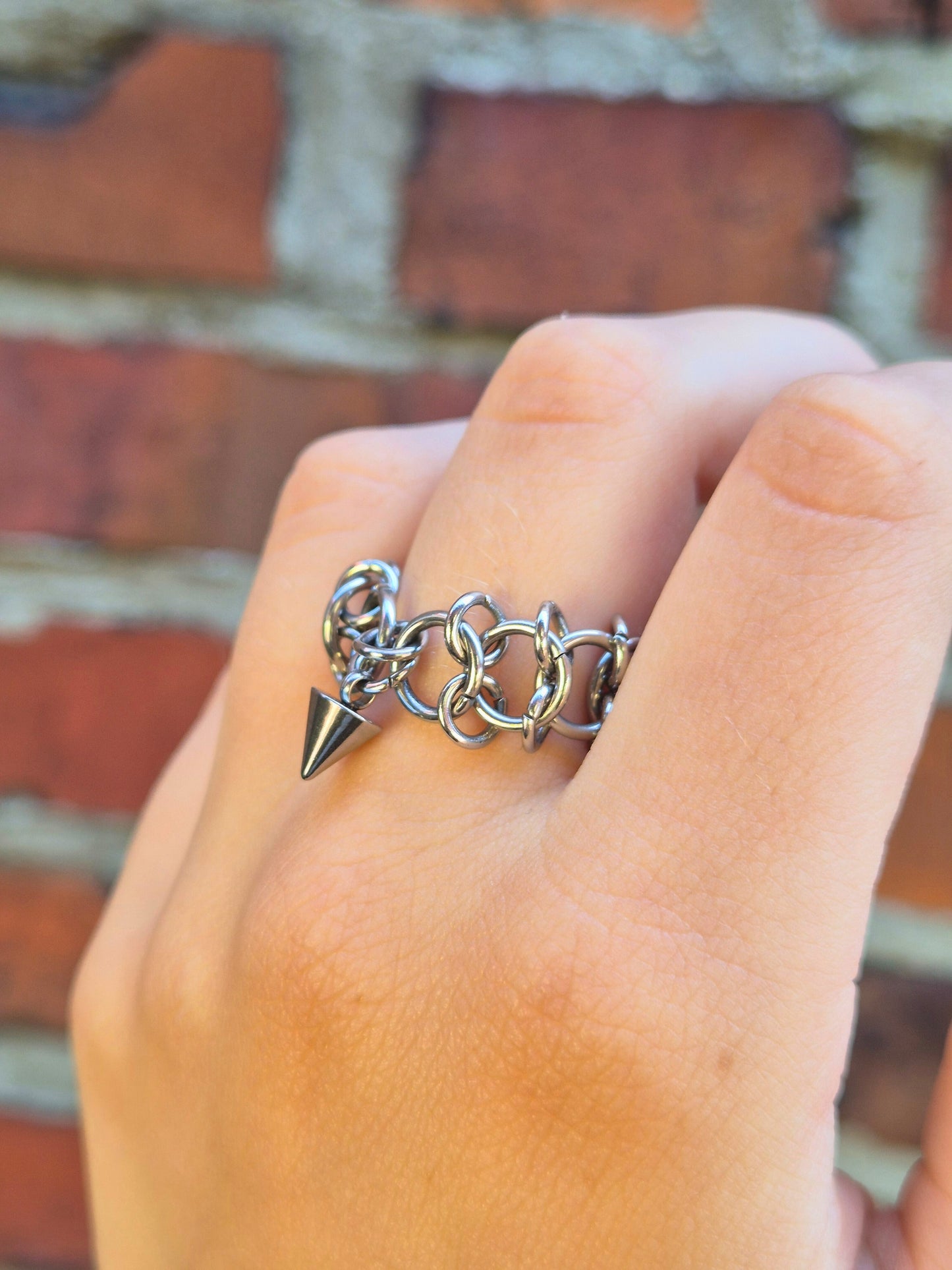Zbliżenie na dłoń z ręcznie wykonaną stalową obrączką w stylu chainmail, ozdobioną metalowym ćwiekiem w kształcie stożków. Pierścień noszony na palcu środkowym. Tłem jest ceglana ściana, która podkreśla surowy i alternatywny styl biżuterii.