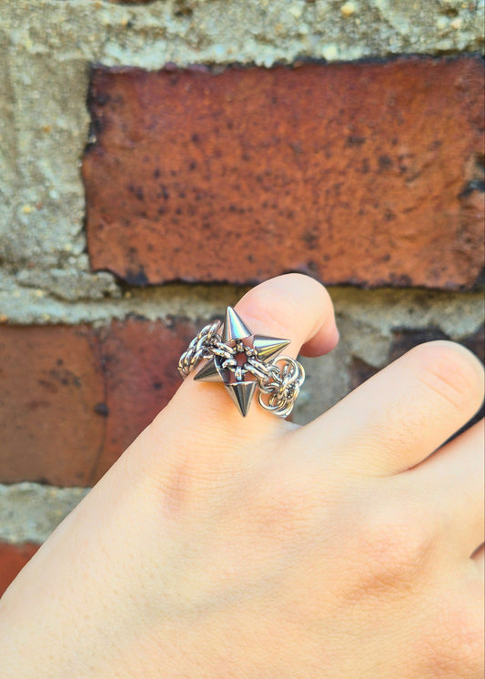Zbliżenie na dłoń z ręcznie wykonaną stalową obrączką w stylu chainmail, ozdobioną metalowymi ćwiekami w kształcie stożków. Pierścień spleciony jest w europejskim wzorze 4 w 1 . Tłem jest ceglana ściana, która podkreśla surowy i alternatywny styl biżuterii.