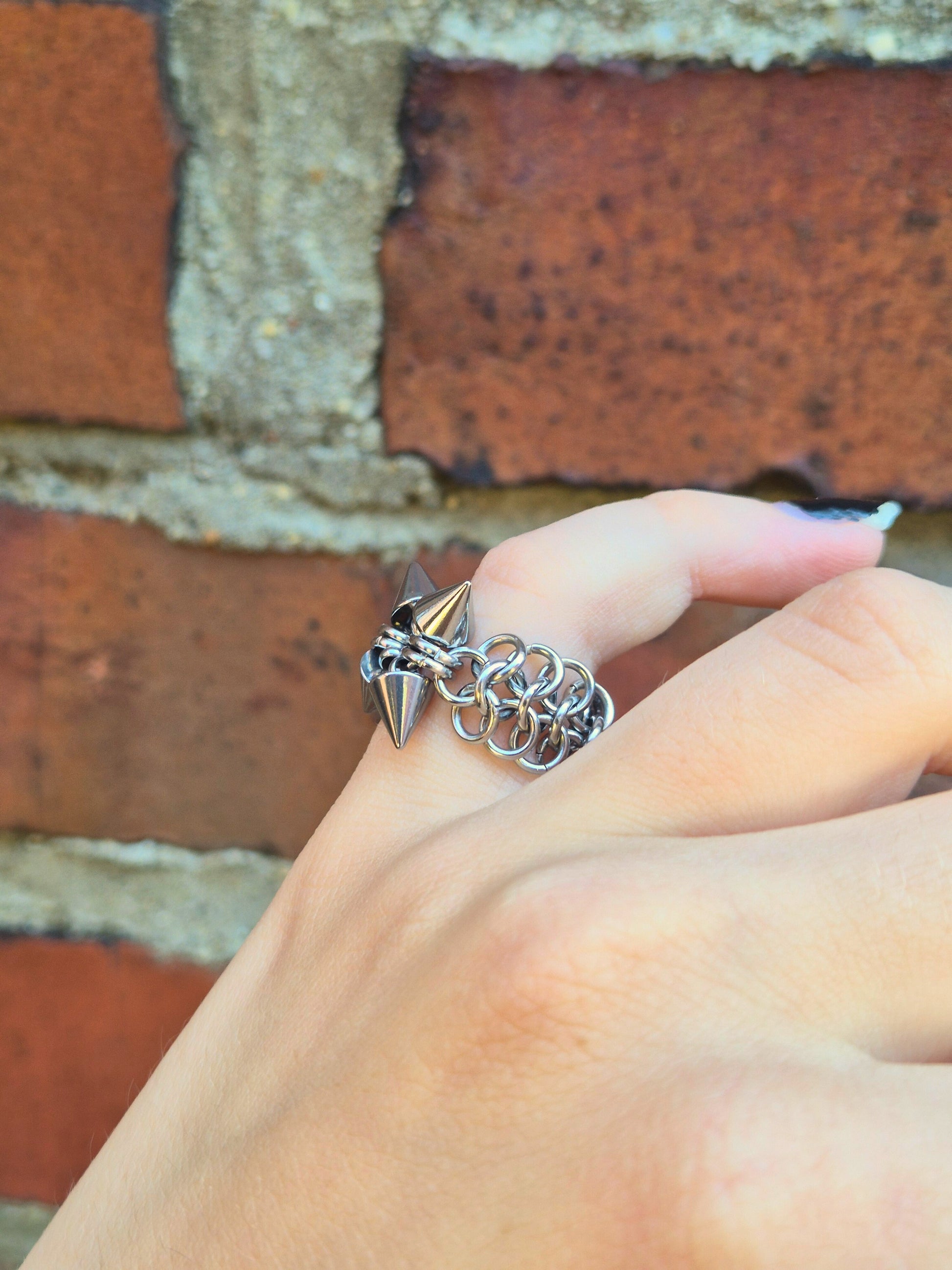 Zbliżenie na dłoń z ręcznie wykonaną stalową obrączką w stylu chainmail, ozdobioną metalowymi ćwiekami w kształcie stożków. Pierścień spleciony jest w europejskim wzorze 4 w 1 i noszony na palcu wskazującym. Tłem jest ceglana ściana, która podkreśla surowy i alternatywny styl biżuterii.