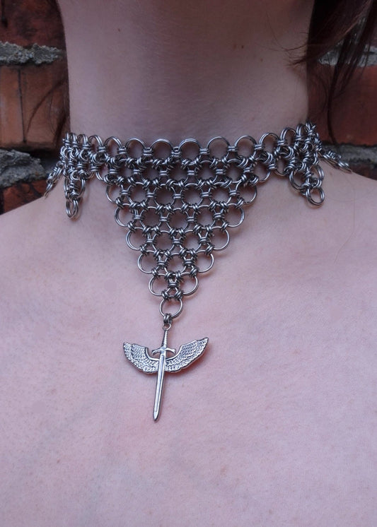 Zbliżenie na choker z kolczugi ze stali nierdzewnej w kształcie odwróconego trójkąta z zawieszką — skrzydlatym mieczem. Biżuteria handmade inspirowana estetyką wojowniczki i magii.